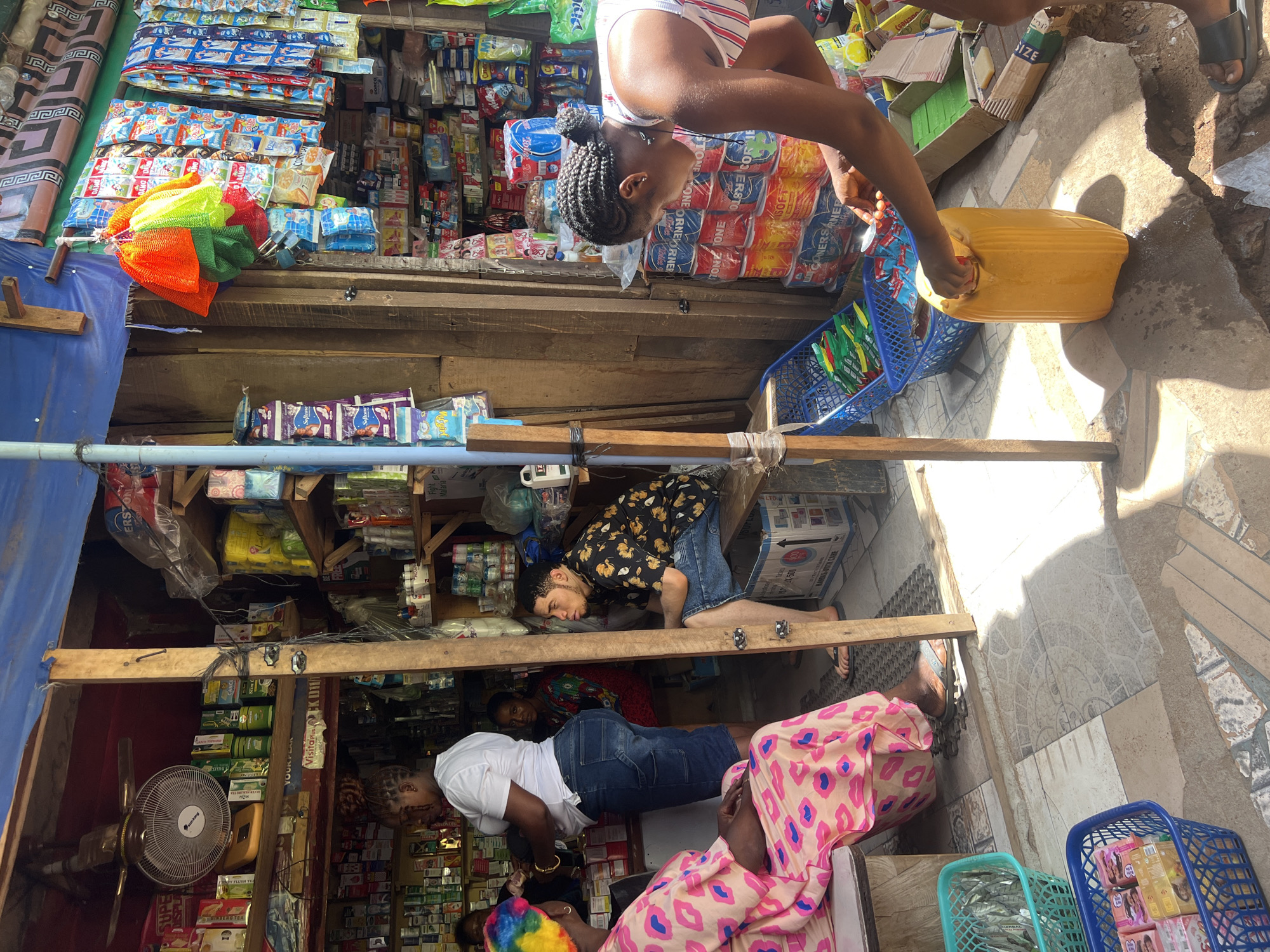 Here's a detailed description of the image:
**Location:** The image is taken in a marketplace environment in Nkpor, Nigeria.
**People:** The image depicts several people, including:
* A young woman in a striped top is bending over and appears to be handling a yellow plastic container.
* Another individual wearing a floral patterned shirt and denim shorts is positioned behind a shelf.
* At least two other people are visible inside the market stall. One is wearing a white shirt and denim shorts. The other is wearing a pink patterned headscarf.
**Objects:**
* A market stall filled with various goods such as packaged food items, toiletries, and other household supplies.
* A metal fan
* Colorful netted scrubbers
* Blue plastic baskets containing various items.
* Wooden shelves filled with a variety of products.
**Actions:**
* The young woman is engaged in some kind of interaction with the yellow container, possibly filling it or opening it.
* People are likely shopping or tending to business in the market stall.
**Environment:** The scene takes place in what seems to be a busy marketplace. There is a wooden stall structure with shelves filled with a variety of products. The ground is paved with tiles.
**Interactions:**
* The most evident interaction is the young woman handling the yellow container.
* Other interactions likely include customers shopping and merchants conducting business in the market stall.