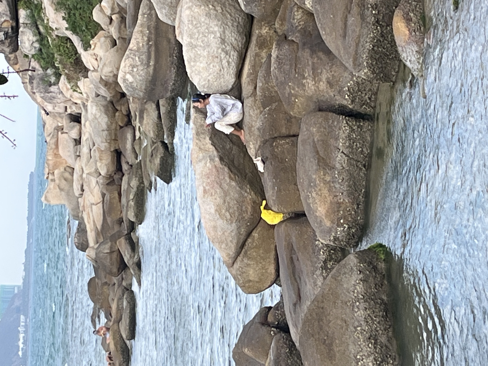 A horizontal medium-shot captures a shoreline scene in Bac Nha Trang Ward, Vietnam. The scene includes a rocky coastline with a person perched amongst the boulders, close to the water's edge. The water is light and looks rough with waves. A woman is climbing on the rocks and is wearing a blue head-band and white clothing. A yellow object is visible near her. Rocks with green foliage are on the left. Other people stand in the water closer to the left. Buildings are visible on the far left horizon.