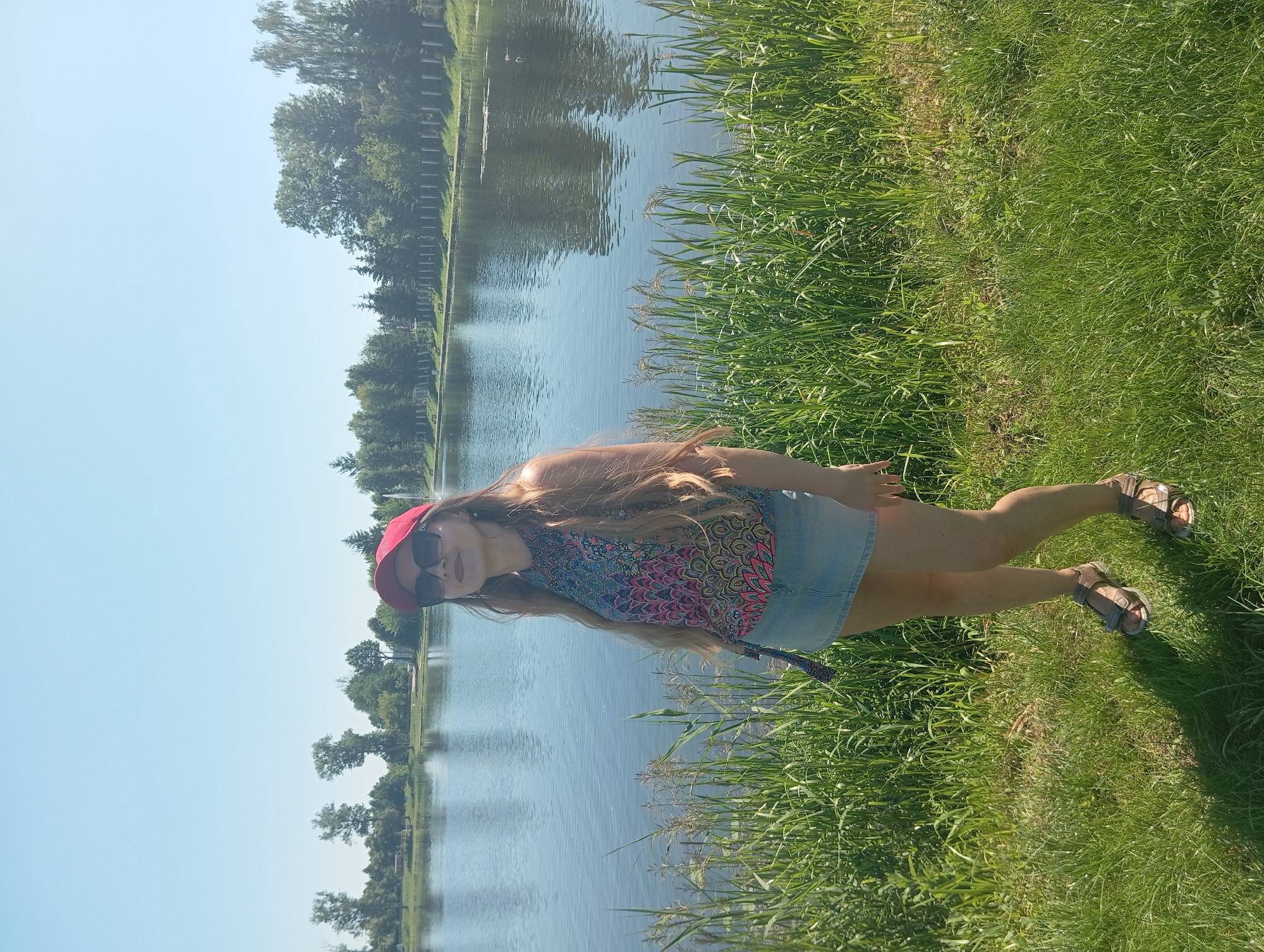 A full-shot, outdoor, medium-light skin toned woman wearing a red baseball cap, sunglasses, a multi-colored sleeveless top, and a light blue denim skirt stands in a grassy area with tall green grass in the foreground. She is facing slightly left with her head tilted back and her mouth open in a smile. Behind her is a large body of water, likely a lake or river, with trees and a fence line along the far bank under a clear blue sky. The scene is set in the city of Wasilków, Poland.