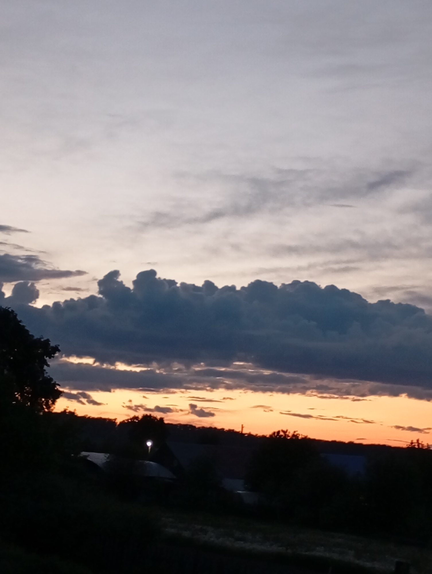 The image shows a landscape at dusk or dawn in Фоки, Russia. The sky is predominantly gray and white with horizontal bands of clouds. A thick layer of dark, billowing clouds is situated below the lighter sky. An orange-hued glow is visible between the dark clouds and the horizon. The foreground is a dark silhouette of trees and possibly buildings, with a single bright light visible amongst the structures. The scene appears calm and still, with no clear indication of people or activity.