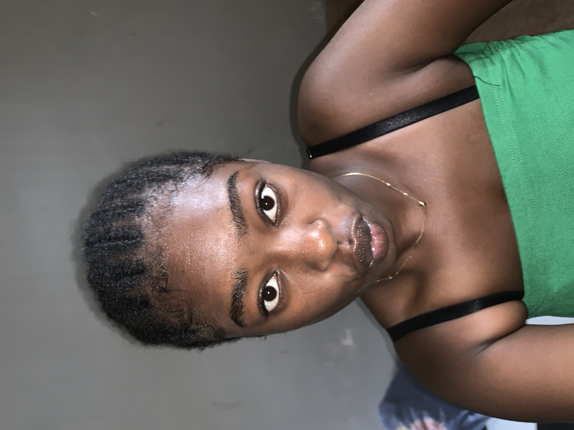 A person with dark skin is facing the camera at an angle. They have short, dark hair and are wearing a green top with black straps. A gold necklace is visible around their neck. The background is a plain, light gray color. There is no indication of a location in Rafin Par, Nigeria.