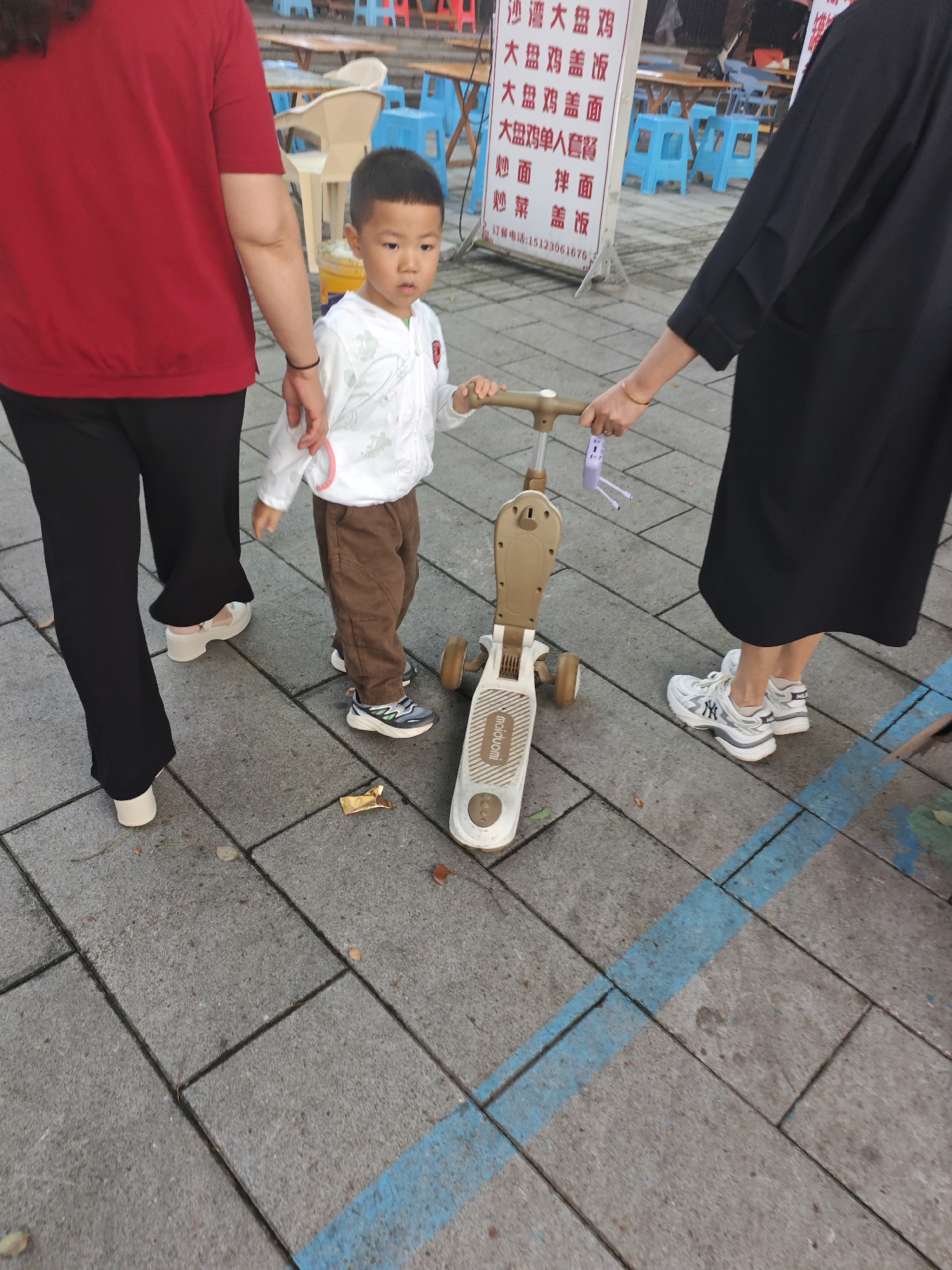 In Chongqing High-tech Zone, China, a young boy, estimated to be between 3 and 5 years old, is positioned in the center of the frame, looking towards the camera. He is wearing a white long-sleeved shirt with light green accents and brown pants, with sneakers. The boy is holding the handlebars of a beige and gold three-wheeled scooter, with a small purple toy attached to the handlebars. He is accompanied by two adults. To his left, an adult wearing a red shirt and black pants is holding the boy's hand. To his right, another adult in a black dress is also holding onto the scooter's handlebars. The scene takes place on a paved walkway with a blue painted line running diagonally across it. In the background, tables and blue chairs are visible, suggesting an outdoor seating area. A sign with Chinese characters is also present in the background. The ground is a concrete paver pattern, with a few scattered fallen leaves and a piece of golden foil wrapper.