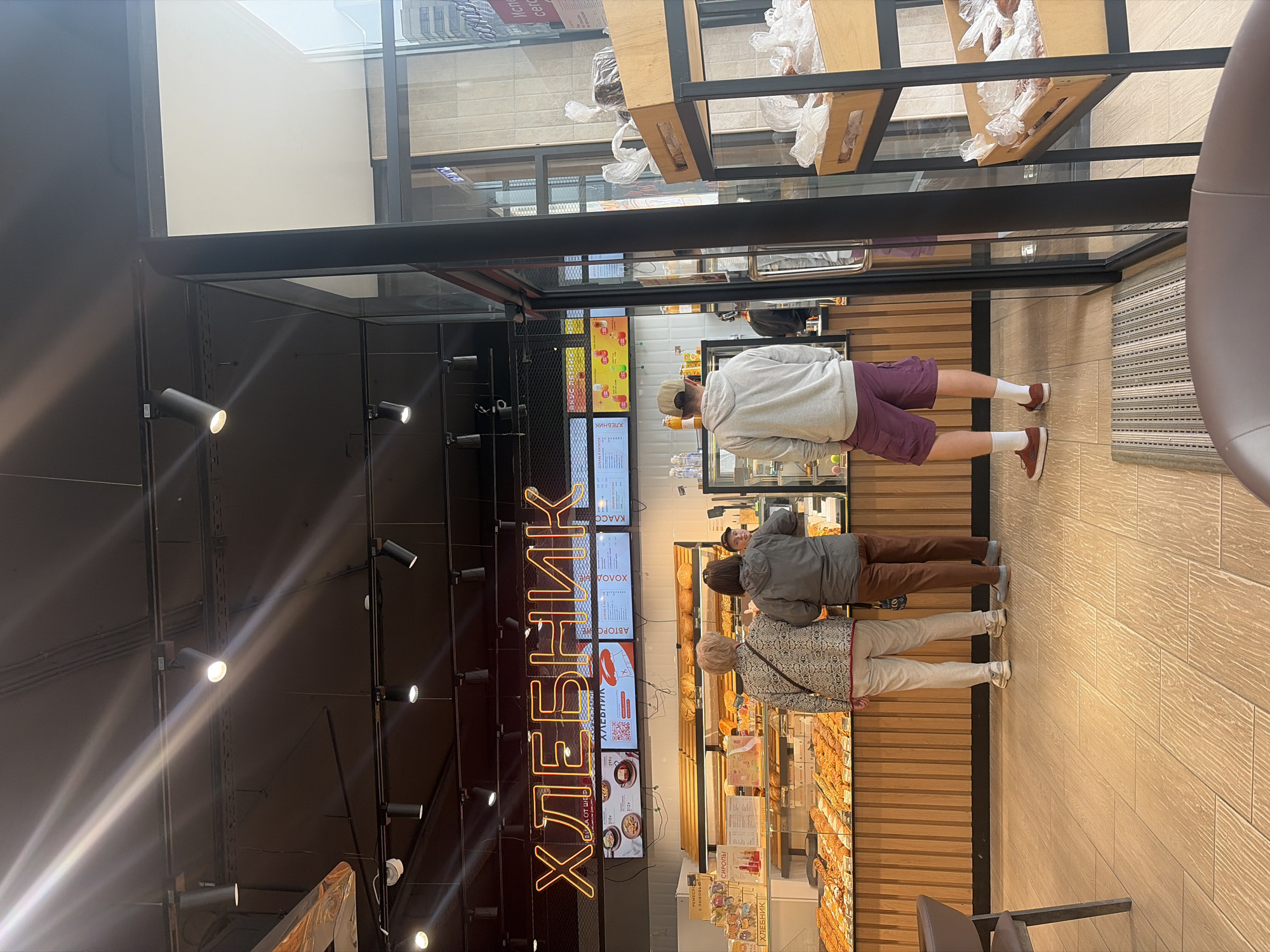 Here is a detailed description of the image:
**Scene:** The image shows the interior of a "ХЛЕБНИК" (Bread Baker) bakery store. There are customers browsing the bakery's display case which holds various pastries and baked goods.
**Location:** This bakery is likely located in Saint Petersburg, Russia, given the brand name ("ХЛЕБНИК") which is a bakery chain prominent in that city.
**People:** Three people are visible in the image. One person in a grey hoodie is standing closest to the bakery staff preparing drinks or coffee. Another person wearing a grey jacket and brown trousers stands behind the hoodie person. Further back is an older woman wearing a patterned jacket and light trousers. They are likely customers selecting pastries.
**Objects:** The bakery counter displays a variety of pastries. The menu board above is visible, which contains prices.
Above the menu board is a sign that reads "ХЛЕБНИК" in large vertical neon letters. Lighting fixtures in the bakery are visible, including spotlights. The bakery has a glass display case to keep food fresh.
**Actions and Interactions:** The people appear to be deciding on their pastries or waiting to order. One of them might be ordering a drink or coffee. The customer in the grey hoodie is holding a bottle of juice.
**Environment:** The bakery has a modern design, with wooden planks covering the lower section of the walls. The flooring looks to be made of wood tiles. The scene suggests a casual setting, typical of a bakery or cafe.