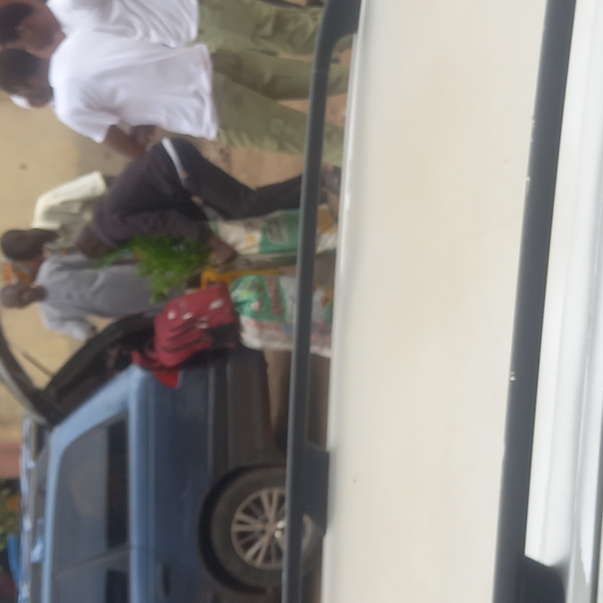 In Sardauna, Nigeria, the image shows several people standing near vehicles. Two individuals wear white shirts and light green trousers. One person wears dark trousers and a dark jacket. Two other individuals wear light-colored shirts. A red bag is visible near sacks of goods. The vehicles are blue and white, with a visible tire on the blue vehicle. The environment appears to be a street or open area.