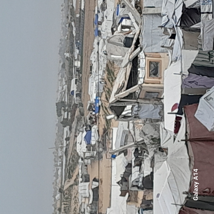 An aerial view displays what appears to be a refugee camp, densely populated with makeshift shelters. A road-like pathway runs through the camp. Buildings are scattered along the upper left part of the image. The tents and structures seem to be made of white, tan, and brown materials. Clothes lines are visible. A general sense of temporary and crowded living conditions is conveyed.