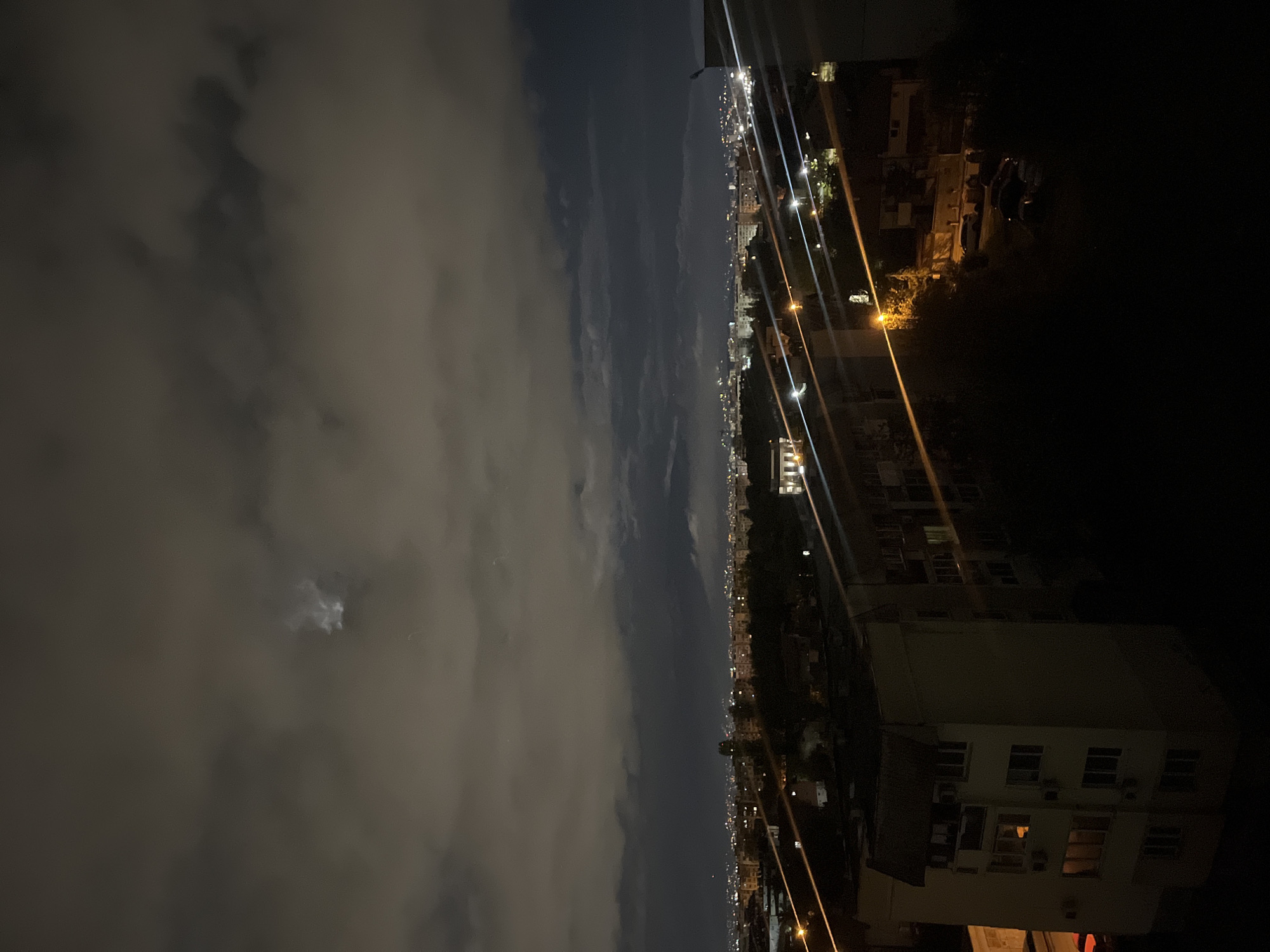 Captured at night, the image presents a panoramic view of Sofia, Bulgaria. Dense clouds occupy the left portion of the frame, punctuated by a faint moon. The cityscape unfolds on the right, illuminated by numerous city lights and street lamps that stretch into the distance. Residential buildings are discernible, their forms shadowed in the prevailing darkness. Overall, the image depicts a cityscape under a clouded night sky.