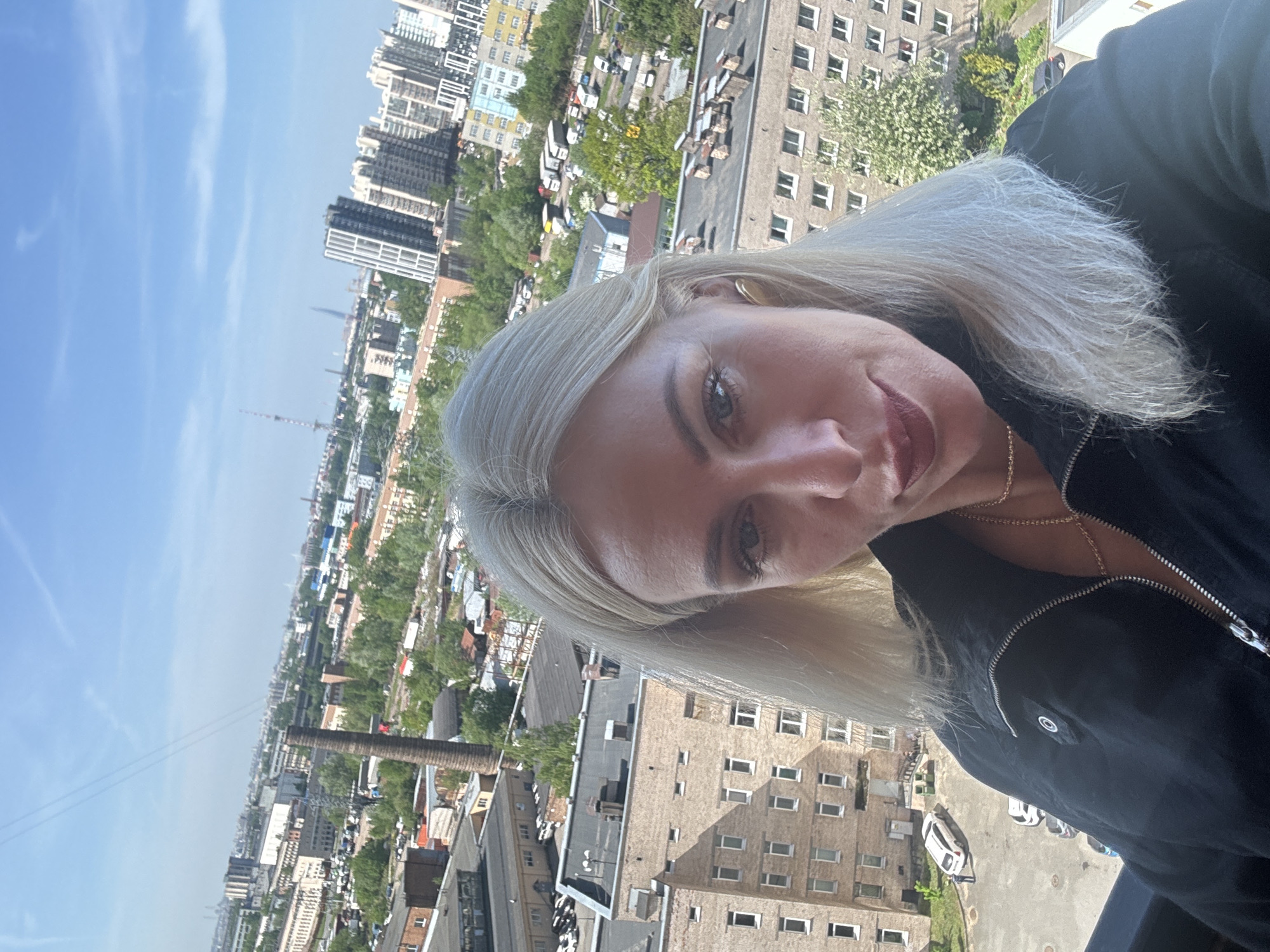 A selfie of a fair-skinned woman with blond hair, wearing a black jacket and gold necklace, is set against an urban backdrop. The city skyline includes buildings of varying heights, some modern high-rises and older brick structures. A brick chimney is visible amongst the buildings. The sky is blue with sparse clouds.