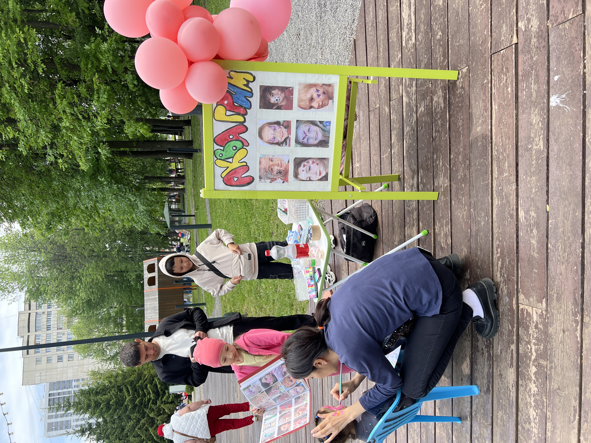 The image shows an outdoor face painting stall, likely in a park in Ufa, Russia, given the urban surroundings and vegetation. A woman, kneeling on a blue plastic chair, is applying face paint to a young child seated in front of her. The child is looking up at the face painting artist. Another child, wearing a pink hat and jacket, is looking at a book of face painting designs being held by an adult standing next to them. A man in a black jacket and white shirt is looking at the child and the book. To the left of them, another person in a hooded sweatshirt stands with their hands on their hips, observing the scene.
Behind the face painter, a small white folding table holds a variety of face paints, brushes, and other materials. A black backpack rests on the ground next to the table. Next to the table is a sign on a lime-green easel displaying sample face painting designs. A cluster of pink balloons is attached to the easel. The surface under the stall is made of wooden planks. In the background, there is a playground structure, trees, and a building.
