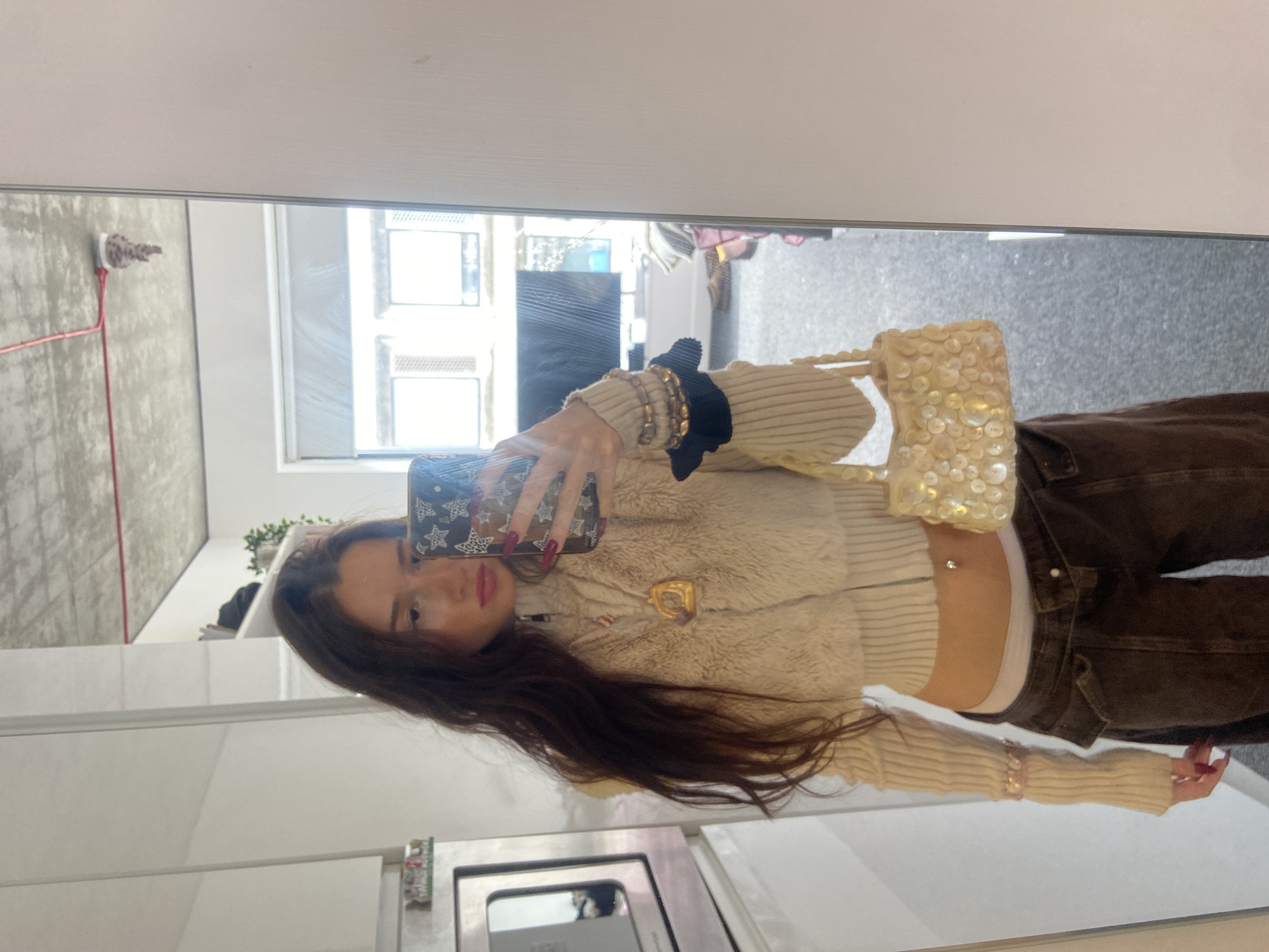 A young woman is captured in a mirror selfie, standing indoors in what appears to be a modern apartment or house. She has long, dark hair and is looking directly into the camera of her smartphone, which has a dark case with a white star pattern. Her lips are adorned with red lipstick, and her fingernails are painted a dark, possibly burgundy, color.
She is wearing a fashionable outfit consisting of a cropped, light-colored top that combines a ribbed knit texture on the sleeves with a fluffy or faux fur material on the body. A white undershirt is visible beneath the cropped top, and she has a silver belly button piercing. On her left wrist, she wears several gold-toned bracelets and a black scrunchie or hair tie. A golden, abstract-shaped brooch or pendant is pinned to her top. She is paired with dark brown or black low-rise cargo pants or jeans. In her right hand, she holds a small, light-colored handbag covered in circular sequins or beads, catching the light.
The background, seen through the mirror's reflection, shows bright daylight streaming through a large window with white frames, suggesting it's daytime. Reflections of what appear to be modern, white kitchen or bathroom cabinetry are visible in the foreground. Further back, there are glimpses of what might be building exteriors or distant objects through the window. A red cord or wire is visible against a textured wall in the upper left background. The overall setting appears clean and contemporary. No discernible text is visible in the image.