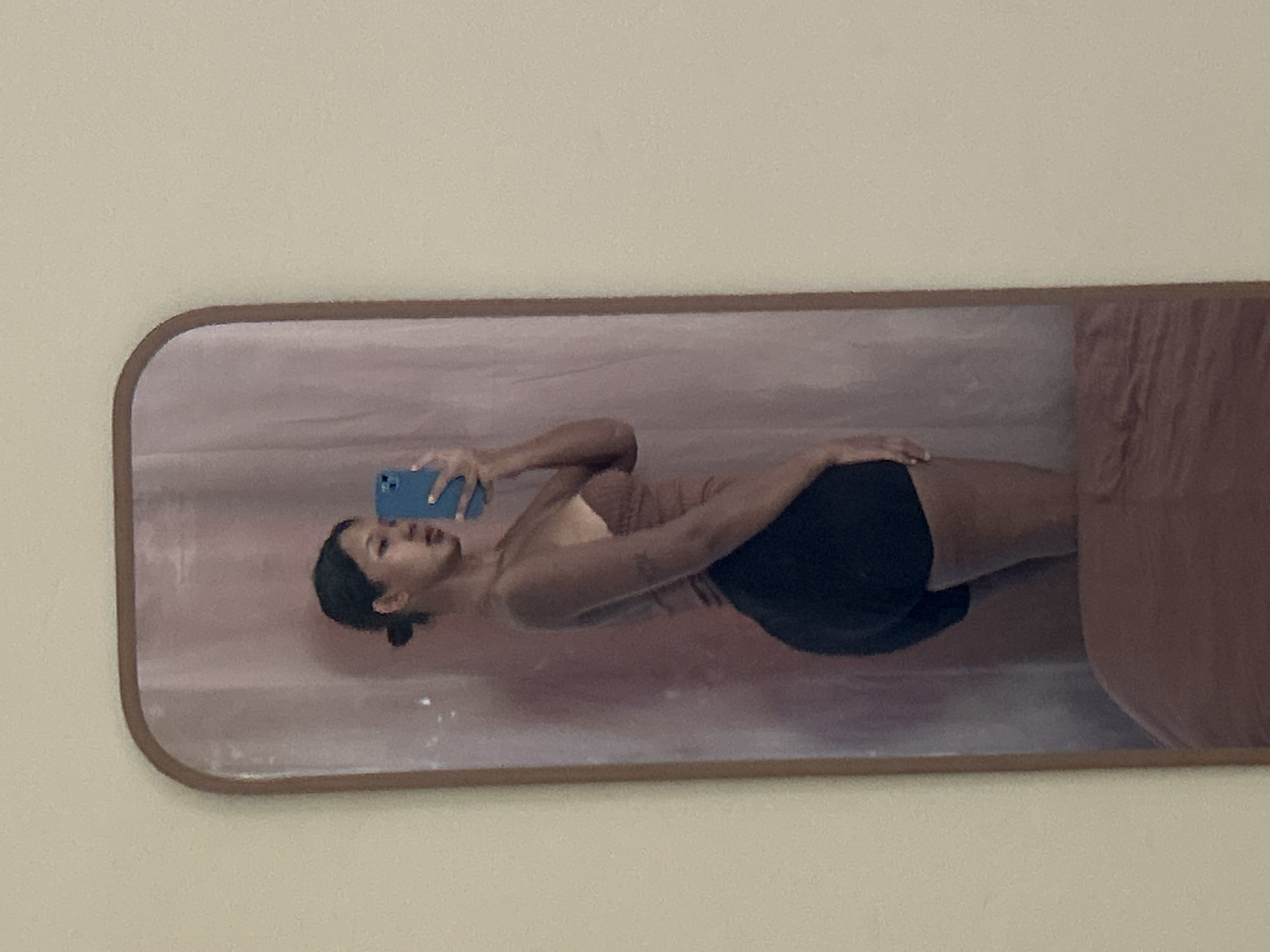 A young woman with dark hair pulled back in a bun stands in front of a mirror, taking a selfie with a blue phone. She is wearing a tan top and black shorts. The mirror is framed with a wooden border and reflects a room with a pale pink curtain in the background and a brown blanket at the bottom. The image is taken from the side, showing the woman's profile and her reflection. The location of this image is not specified in the provided context.