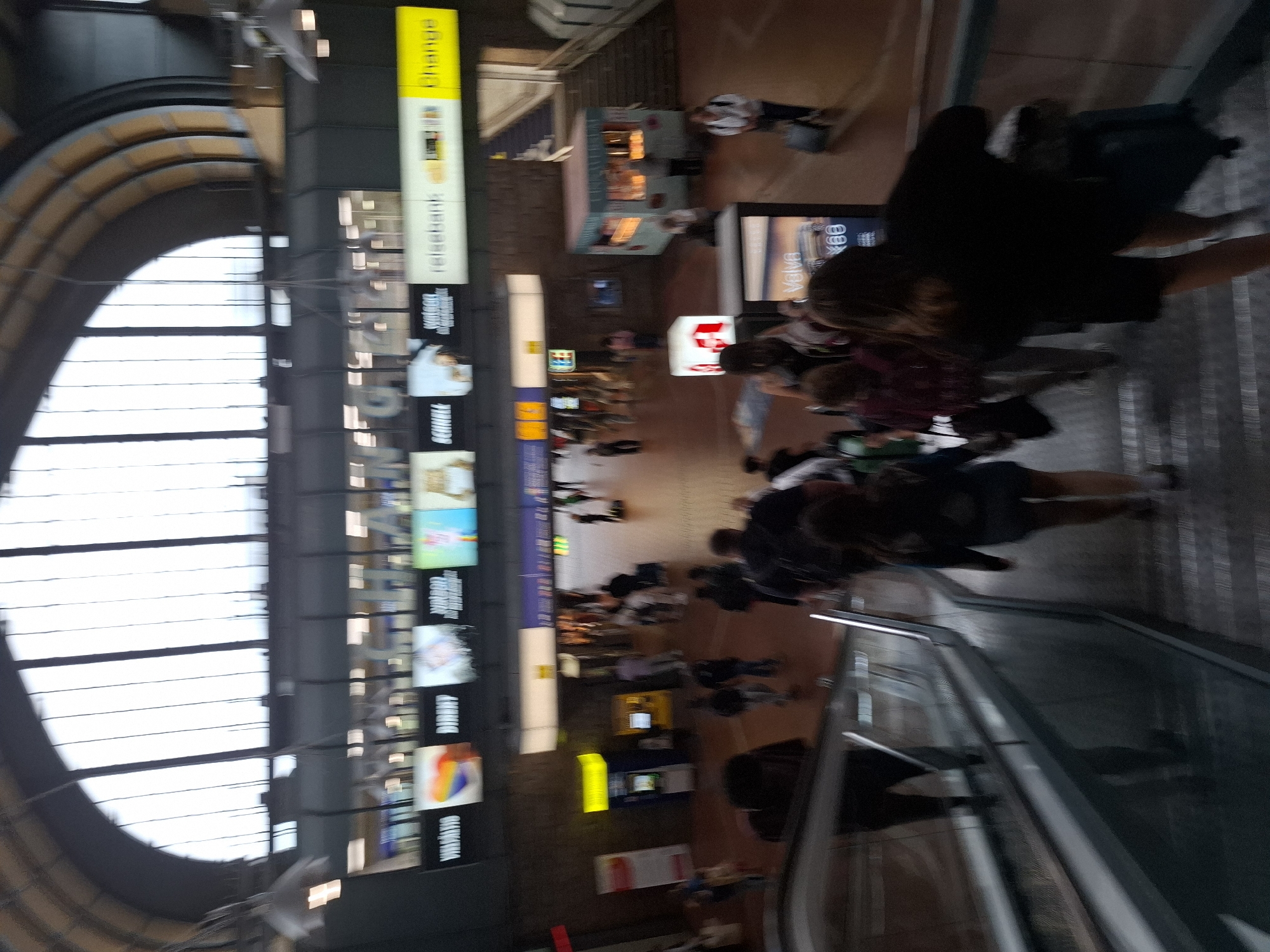 This image depicts the interior of Hamburg Hauptbahnhof, a major railway station in Hamburg, Germany. The scene is bustling with people moving about on escalators, stairs, and the concourse. In the foreground, an escalator is visible, with several individuals ascending and descending. The architecture of the station is characterized by a large, arched glass ceiling, allowing natural light to illuminate the space. Several advertisements are displayed on illuminated panels above the concourse. To the right, a small kiosk or shop is present, with people inside. Further in the background, a staircase leads to an upper level. The overall impression is one of a busy transportation hub.