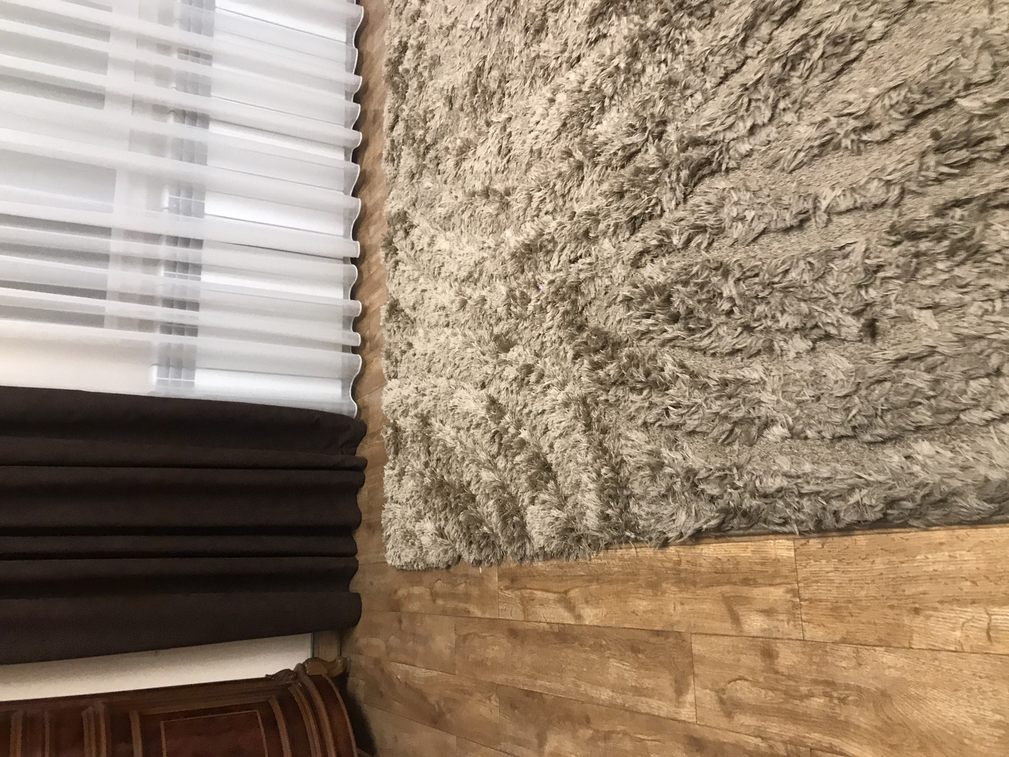 The image shows a section of a room. On the left, there's a window covered by horizontally striped blinds, part of which are white and translucent, while the lower section is a dark brown pleated fabric. To the right of the window is a large beige, shaggy rug that is partially visible. Below the rug, the floor is made of light brown wood planks. The bottom left corner reveals a portion of a dark-stained wooden furniture piece, possibly a headboard or decorative element. The image provides no information to suggest its location in Yerevan, Armenia, or anywhere else. No people, interactions, or actions are depicted.