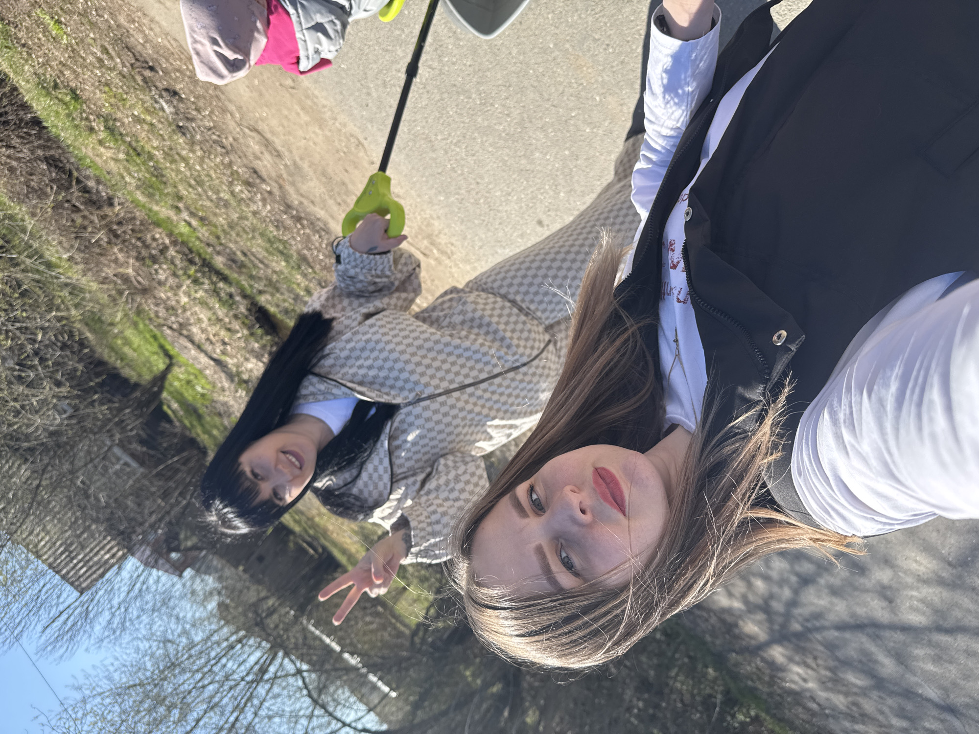 The image shows two women taking a selfie outdoors on a paved surface resembling a path or road. One woman, positioned closer to the camera, has long brown hair and is wearing a white shirt under a black vest. The other woman, standing slightly behind, has long black hair and is wearing a light-colored, patterned jacket. She is holding a green handle, likely part of a child's stroller or tricycle, and making a peace sign with her hand. Part of a child's pink and grey jacket is visible in the upper left corner, along with a white container, seemingly attached to the tricycle or stroller. The background includes trees without leaves and what appears to be the edge of a building. The location is specified as Zav'yalovo, Russia, although specific landmarks are not discernible in the frame.