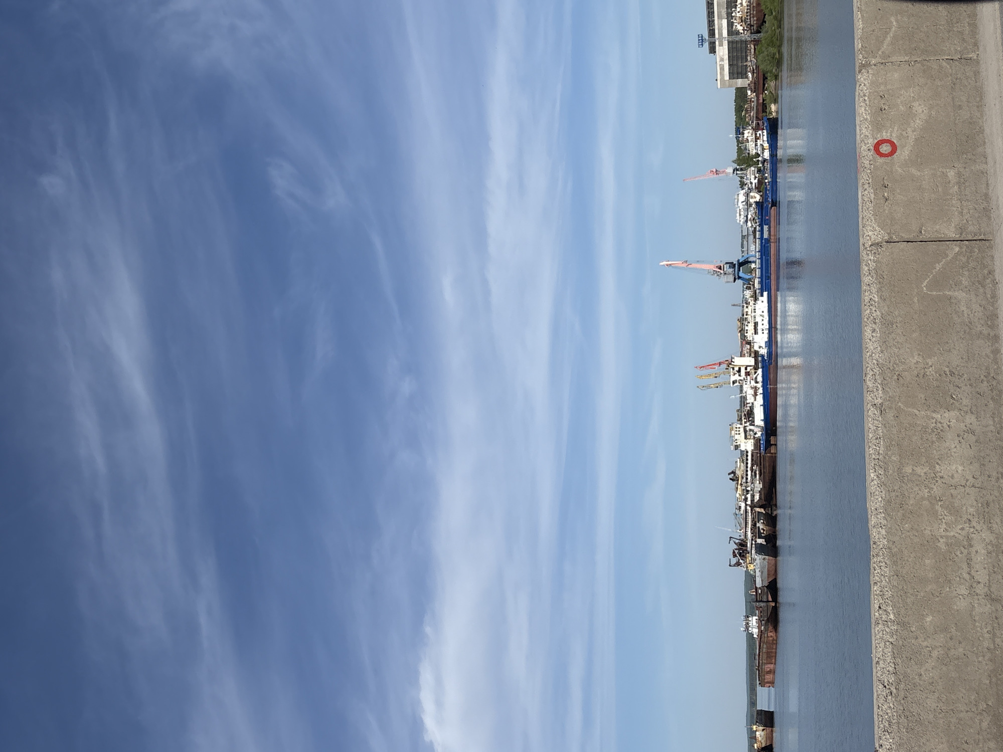 The image shows a view of a waterfront, likely a port or harbor, in Chaikovsky, Russia. There are several ships and barges docked along the water's edge, with various cranes and industrial structures visible in the background. The sky is blue with wispy clouds. A concrete wall or embankment runs along the right side of the frame, with a red circle painted on it. The photo appears to have been taken on a clear, sunny day.