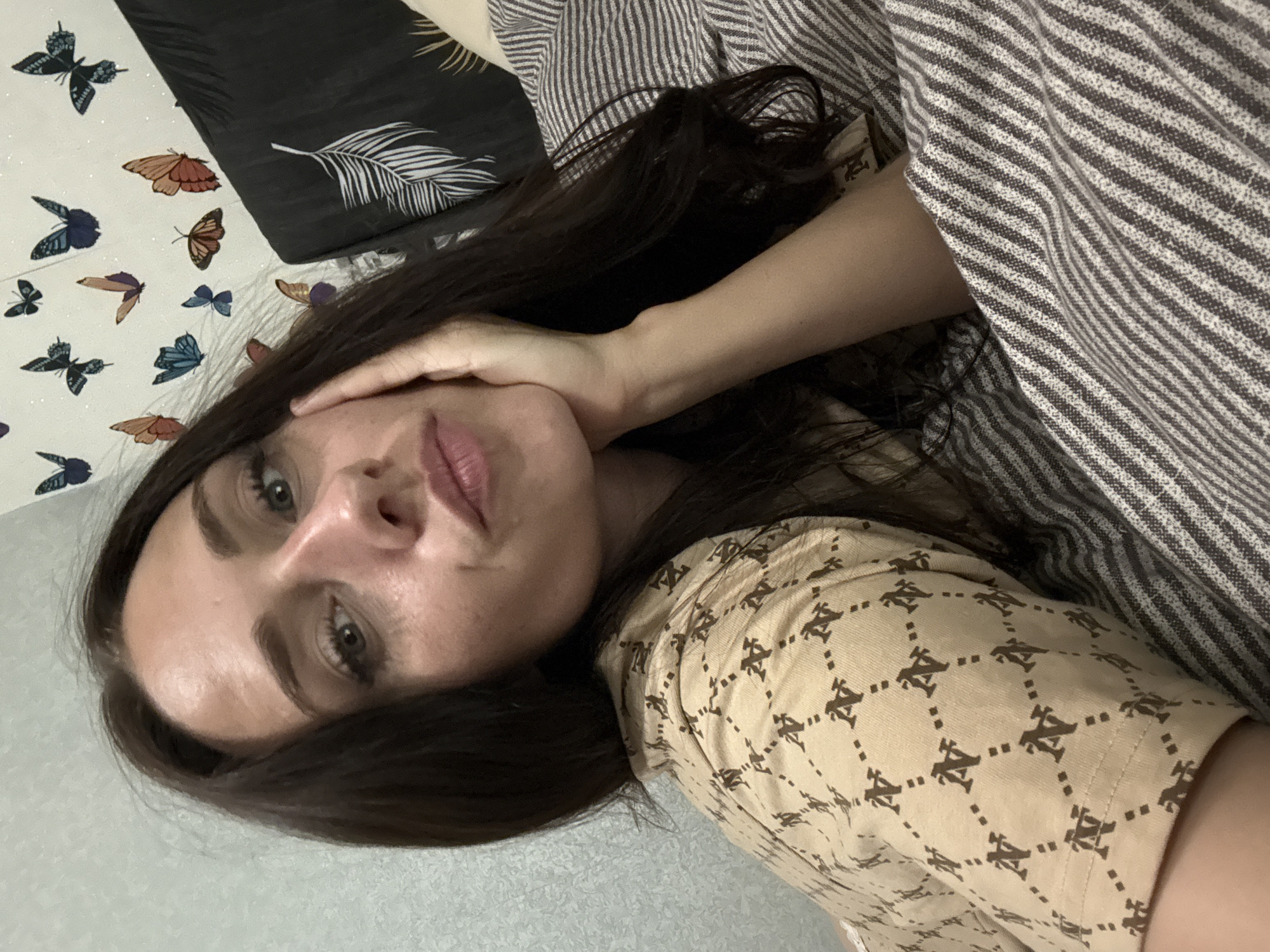 In this close-up selfie taken indoors, a woman with long, dark hair rests her head on her hand, looking towards the camera with her lips slightly pursed. She wears a tan shirt adorned with a repeating pattern of interconnected shapes containing the letter "N." The background features a wall decorated with a collage of butterfly decals in various colors and sizes. To the right of the woman, a pillow with a black cover featuring a white feather pattern is visible, partially obscured by a striped blanket. The overall impression is that of a casual, personal moment captured in a domestic setting, possibly a bedroom. The location in Saint Petersburg, Russia, is not discernible from the visual cues in the image.