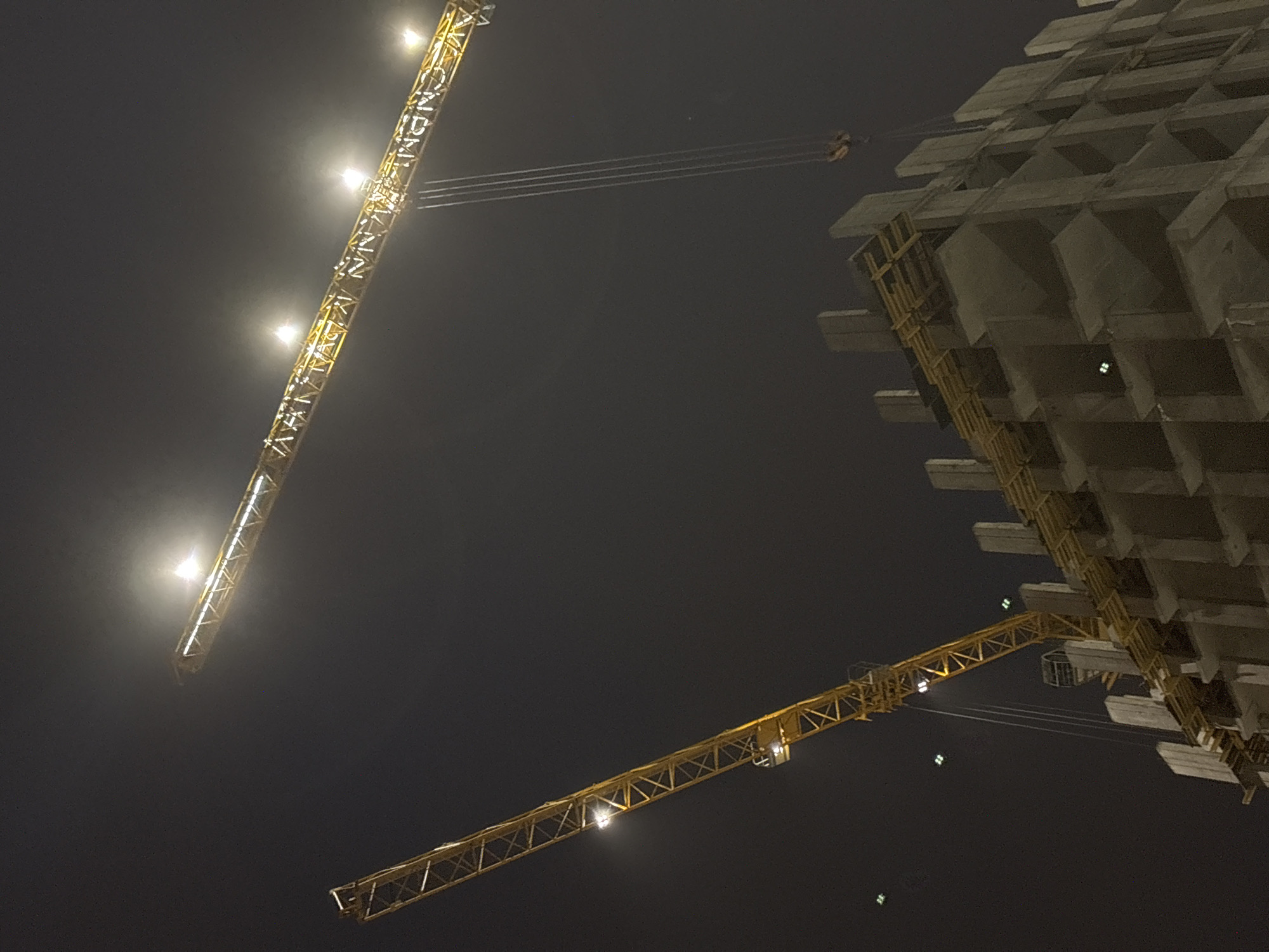 The image depicts a construction site at night, likely in Perm, Russia, given the request for location. A large concrete building is under construction, with two tower cranes visible against a dark sky. The cranes are illuminated with lights, and their arms extend towards the building.