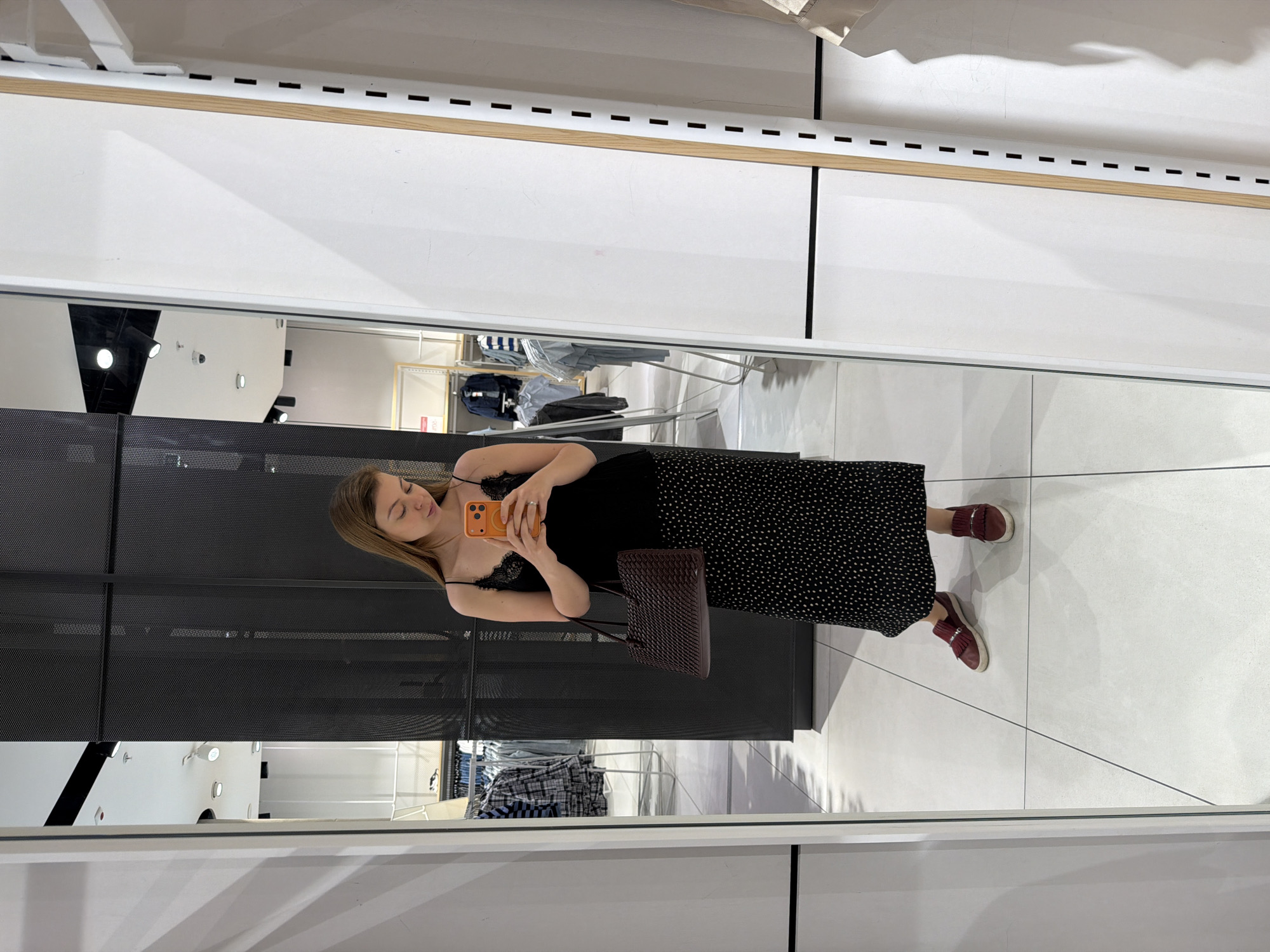 The image shows a woman taking a selfie in a mirror inside a store, potentially in Khimki, Russia, based on visual context. She is wearing a black, spaghetti-strap top with lace details and a long, black and white polka-dot skirt. She has brown hair and is holding an orange smartphone. She is holding a brown woven handbag. She is wearing burgundy loafers. The mirror reflects the interior of the store, including clothing racks with various garments. The store appears to have a modern design with white walls and a dark, perforated metal display structure. The floor is made of white tiles.