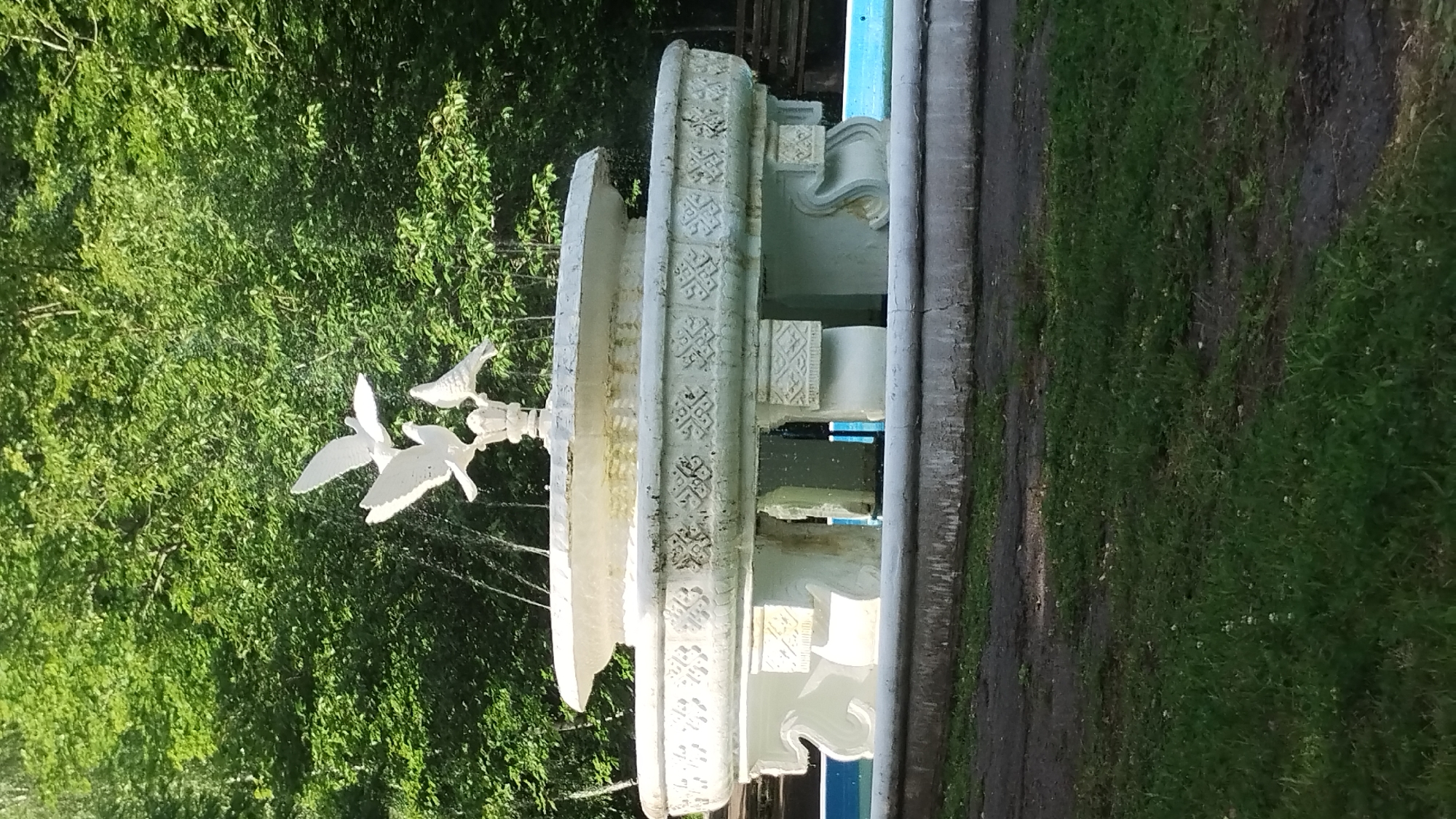 The image shows a white, multi-tiered fountain in a park setting, featuring white dove sculptures at the top, with water spraying from the fountain. The fountain has intricate carvings along its edge and is supported by pillars. In the background, lush green trees form a dense canopy. The foreground consists of grass and a paved path. The fountain appears to be located in a public space, possibly a park or a square, in the city of Syniak, Ukraine.