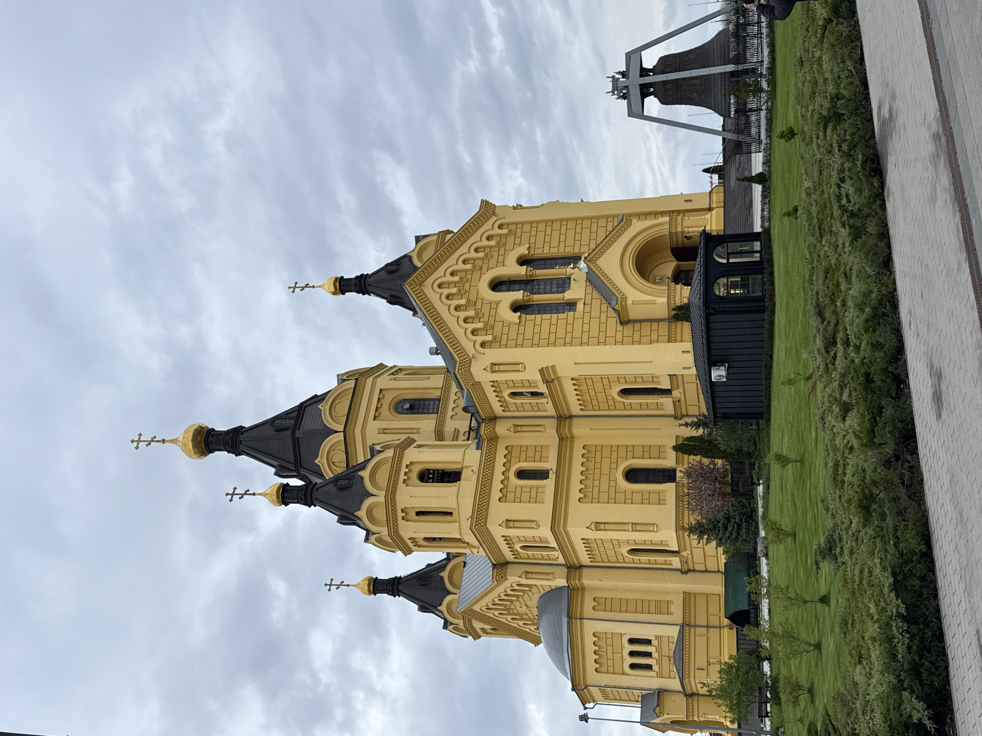 The image shows the Alexander Nevsky Cathedral in Nizhny Novgorod, Russia. The cathedral is yellow with black spires topped with golden domes and crosses. A large bell hangs on a metal structure near the cathedral. A green lawn and low shrubs surround the building, and a gray paved walkway is on the right side of the frame. The sky is cloudy and gray.