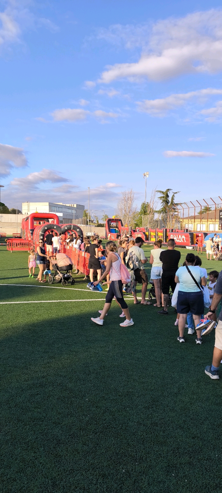 The image captures an outdoor event on a green artificial turf field, likely a sports field, under a bright blue sky with scattered white clouds. In the background, several large inflatable structures, predominantly red and black, are set up, resembling obstacle courses or play areas. A line of people, adults and children, are queuing for one of the attractions, with a red barrier separating them from the field.
Several individuals are visible in the foreground and middle ground, interacting with each other or walking. One woman in a pink tank top and black capri leggings is walking towards the right, carrying a pink bag over her shoulder. Another woman in a light blue t-shirt and dark shorts is standing facing left. A man with tattoos on his leg and wearing a black t-shirt and shorts is also visible. Children are present, some with adults and one in a stroller.
In the middle ground, a red inflatable structure shaped like a vehicle with large black tires is prominent. Further to the right, another similar inflatable structure is visible. Beyond the inflatables, there are buildings and a fence line, suggesting an urban or semi-urban setting. A lamppost is also present. The overall scene depicts a lively outdoor recreational event, possibly a festival or fair, taking place in Ripollet, Spain, given the context.