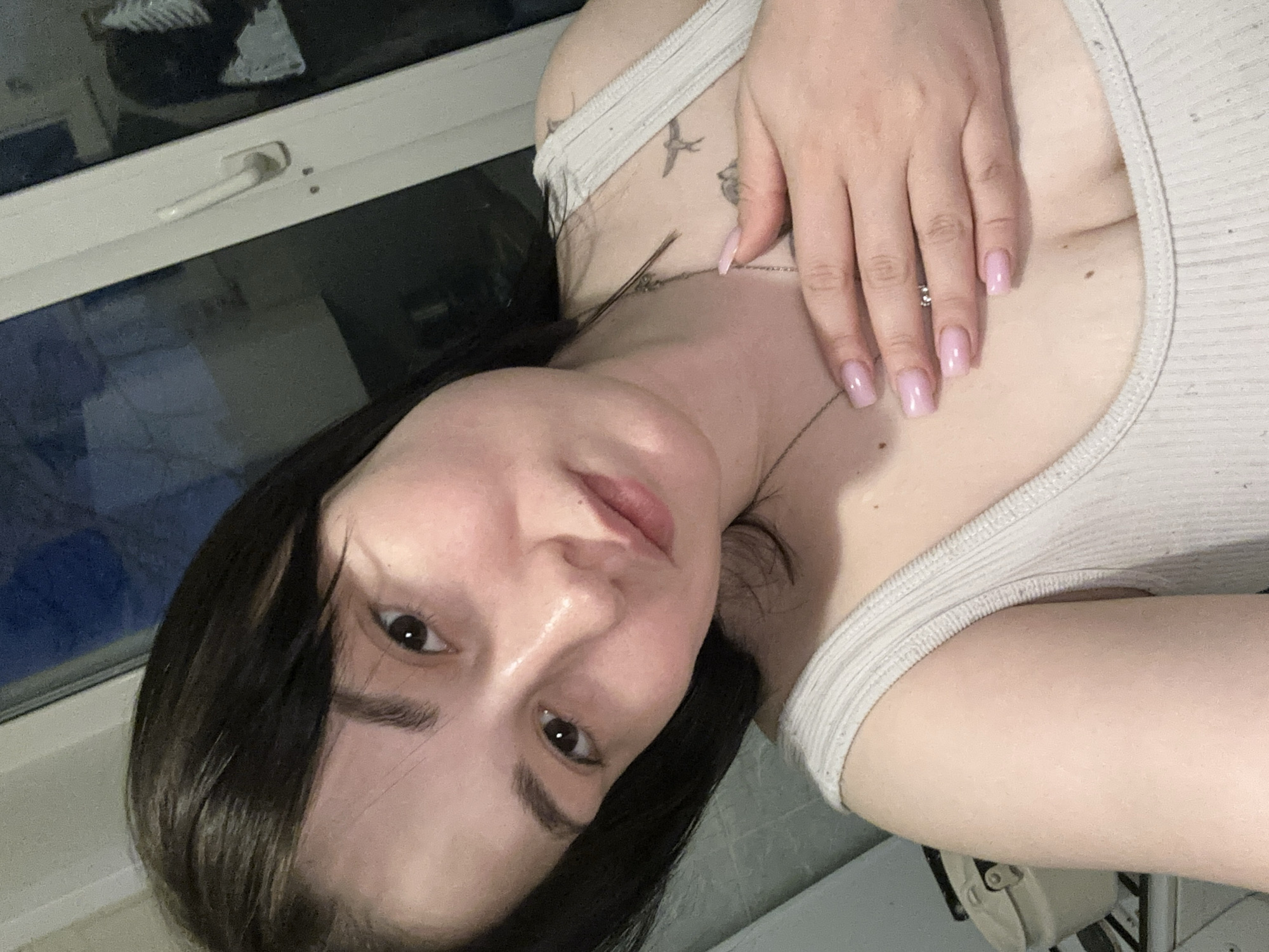 A portrait-oriented selfie captures a young woman indoors in Chaikovsky, Russia, looking directly at the camera with a neutral to soft expression. She has dark hair, fair skin, and dark eyes. She is wearing a light-colored, possibly off-white or beige, ribbed tank top and a thin silver necklace. Her right hand, adorned with light pink square-shaped nails and a small ring on her ring finger, rests gently on her upper chest. Several small, delicate tattoo outlines, appearing to be birds, are visible on her upper chest and shoulder area.
The woman is positioned at an angle, suggesting she is lying down or leaning back against a surface. In the background, a modern window with a white frame and handle is visible. The window glass reflects the dark exterior, indicating it is likely nighttime or late evening, along with indistinct reflections of the room's interior. The lighting on her face and upper body is soft and appears to be artificial. No discernible text is visible in the image.