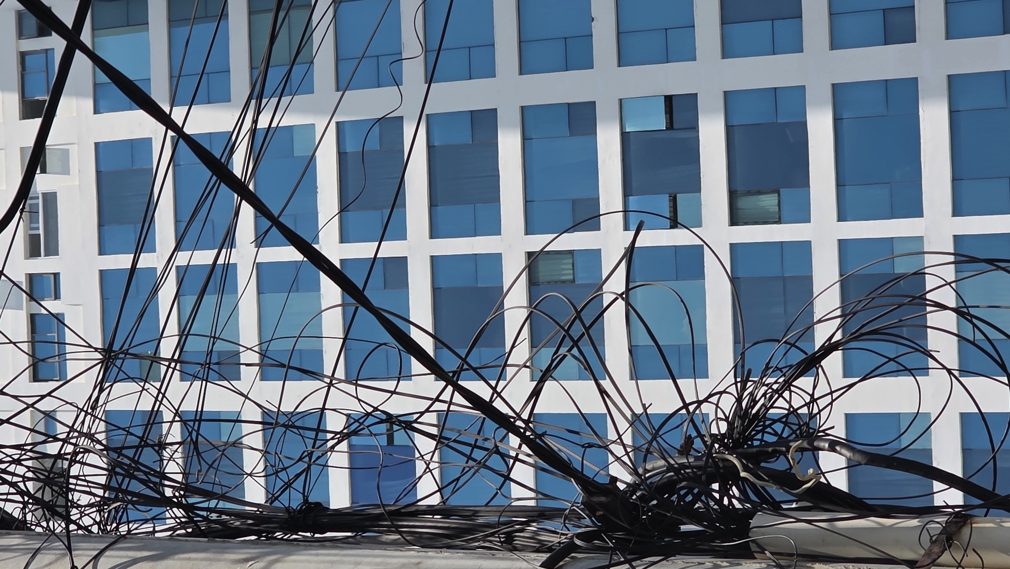 A tangle of black wires is draped across the foreground, obscuring a building with a white grid facade and blue, reflective windows. The wires are of varying thicknesses and appear haphazardly arranged. The building, likely in Bắc Nha Trang, Vietnam, due to the prompt, is the primary background element, with its modern, geometric design contrasting with the chaotic wires. The wires are resting on a white surface, potentially a rooftop or ledge. There are no people visible in the image.