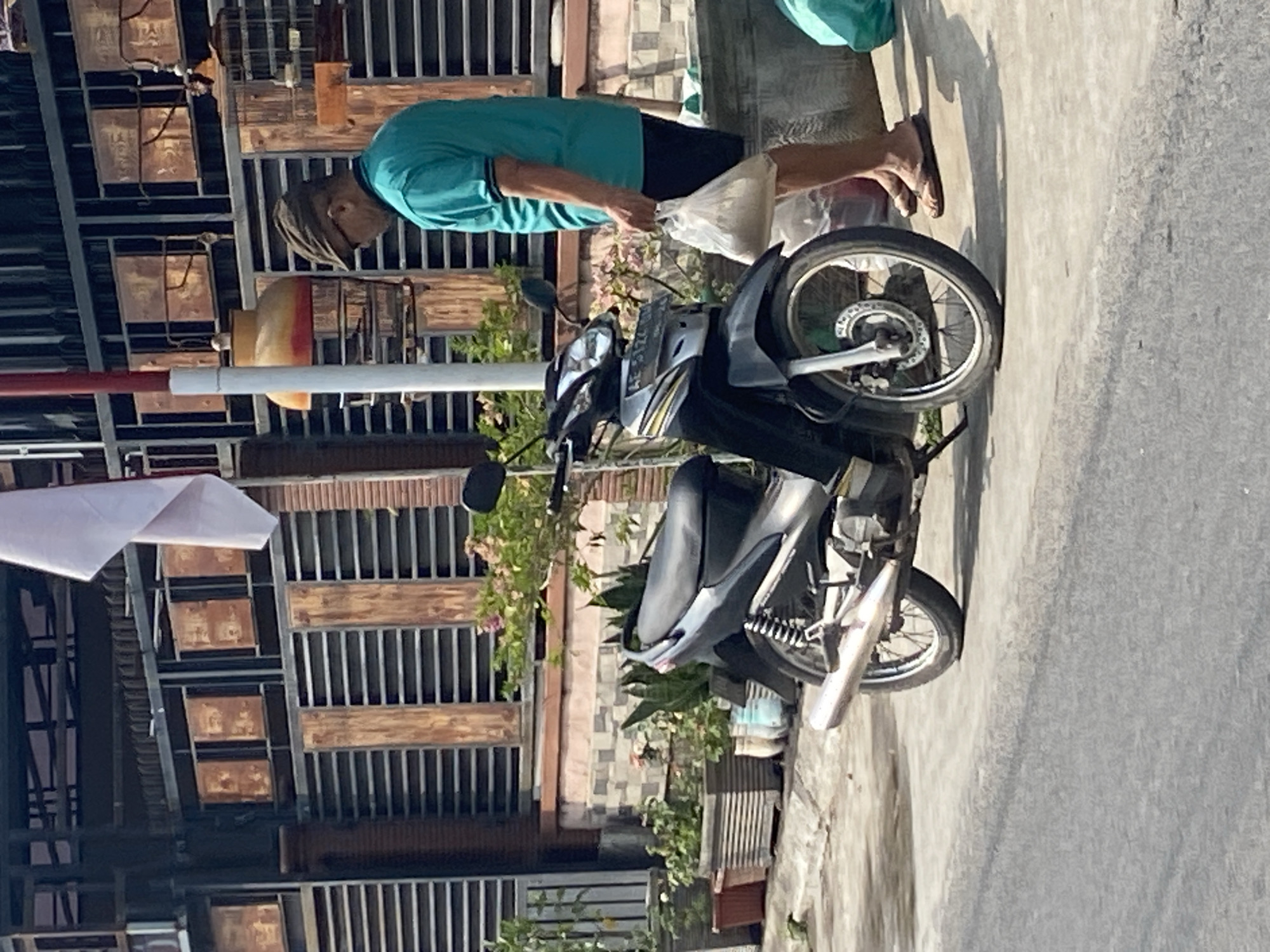 A man wearing a cap and a teal shirt is standing near a motorcycle parked on a sidewalk. He is holding a translucent plastic bag containing light-colored contents. The motorcycle is black and silver with visible front and rear wheels, a muffler, and a license plate. In the background, there is a building with a decorative facade made of wood and metal, along with potted plants and a birdcage. The scene appears to be in an urban setting, likely in Kraton, Indonesia, given the architectural style and the presence of common street elements. The lighting suggests it is daytime.
