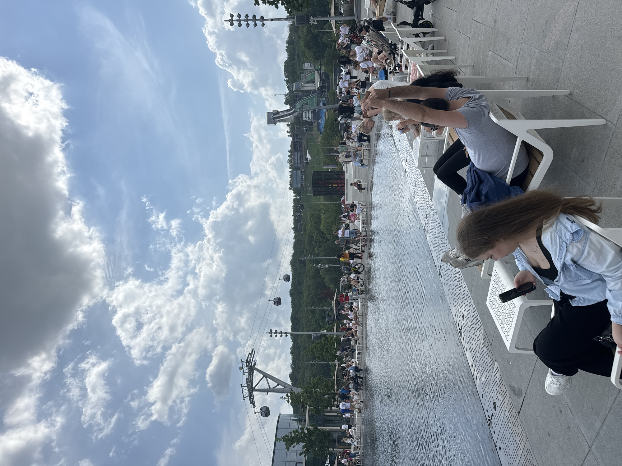 The image shows a group of people sitting in white chairs along a long, narrow body of water, possibly a decorative pool or fountain, in what appears to be a public space. Some people are seated with their feet in the water, while others are looking at their phones or interacting with each other. In the background, there is a forested area and what looks like a ski jump. There are also cable cars visible. The sky is mostly cloudy. Based on these features, the location is likely Zaryadye Park in Moscow, Russia.