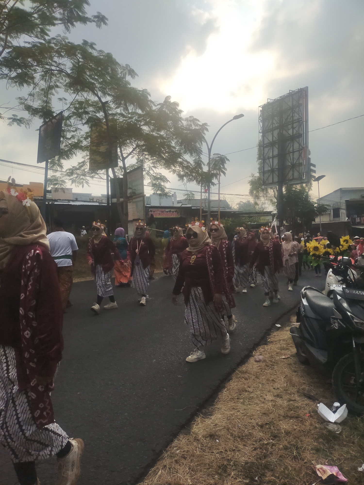 The image depicts a street procession in Tuwel, Indonesia, likely a cultural event or parade. A group of individuals, predominantly women, are walking down the asphalt road. They are dressed in traditional attire, including batik skirts and maroon tops, many adorned with headbands featuring floral or decorative elements. Some are wearing sunglasses. In the background, buildings and trees line the street. A prominent billboard structure stands on the right side of the road, and a motorcycle is parked on the side. The sky is overcast with bright sunlight breaking through. The overall scene suggests a vibrant community gathering under a slightly hazy sky.