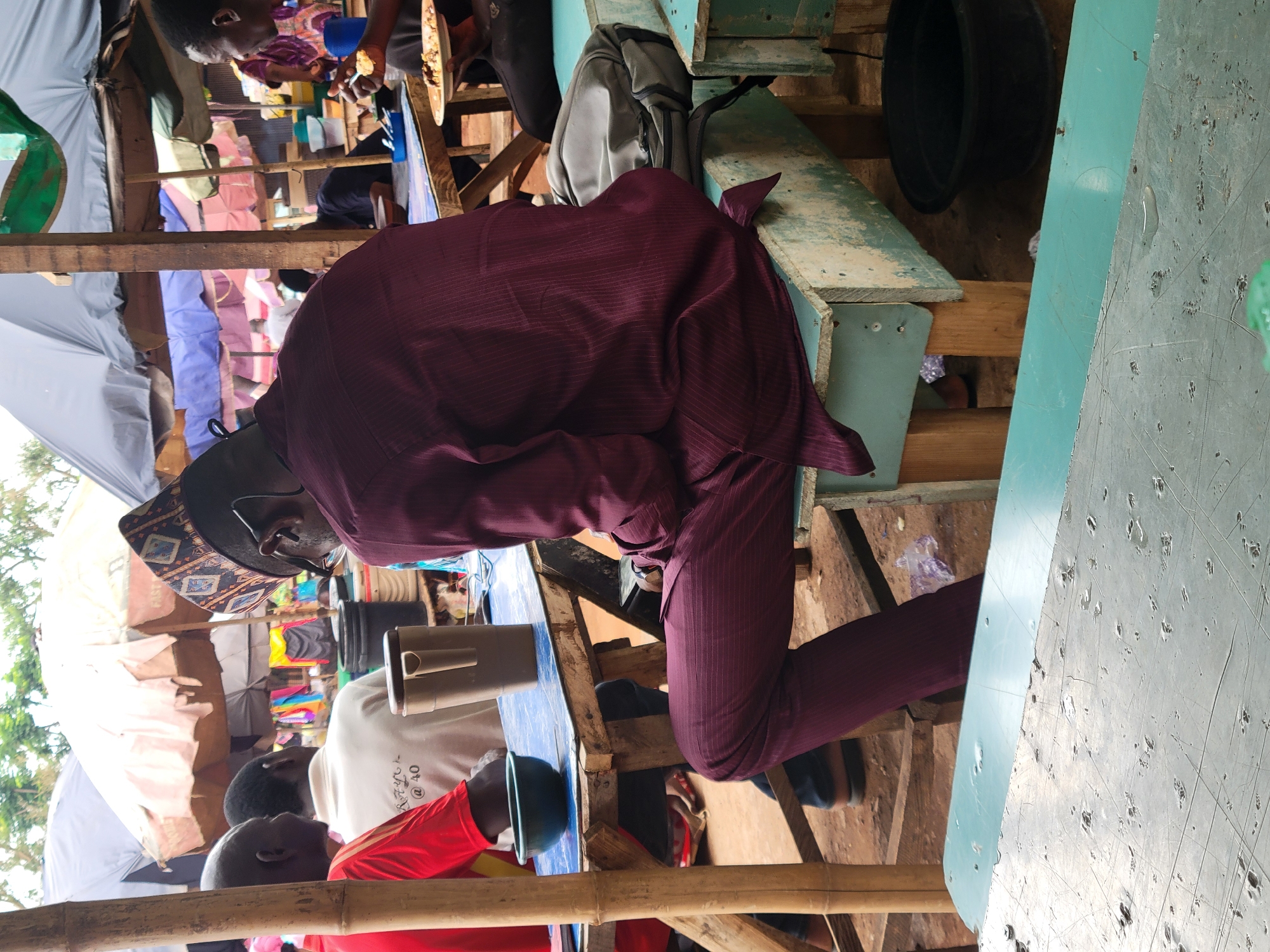 An image of a man in a maroon suit sitting at a table among other people. The man is wearing a traditional patterned hat and glasses. There are several other individuals at the table with him, with a person in a red shirt visible in the foreground. The scene appears to be in an outdoor or semi-outdoor setting, possibly a market or food stall. Colorful clothing items are visible in the background, suggesting a marketplace. The table and seating appear to be somewhat makeshift and rustic. The environment suggests a bustling and crowded location. While the specific location in Abuja, Nigeria cannot be determined from the image alone, the overall context points to a public space within the city.