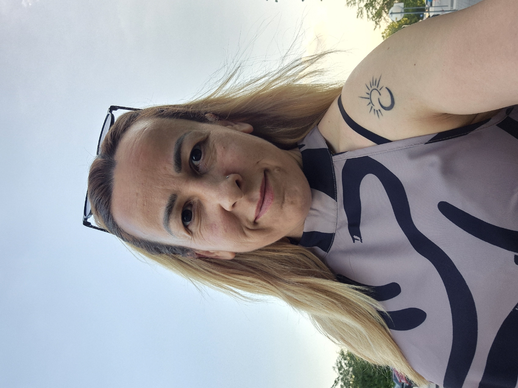 A female subject is depicted in a front-facing outdoor selfie, smiling faintly. She has light blonde hair, worn long and parted on the left, partially obscuring her left shoulder. Glasses are positioned atop her head. A small silver stud piercing is visible on her left nostril. Her right upper arm features a tattoo consisting of a stylized black sun with rays emanating from a crescent moon. She wears a sleeveless top with a high neckline, in a light grey-beige color with a prominent abstract pattern of dark grey-black curvilinear shapes. A black strap, likely from an undergarment, is visible on her right shoulder. The background features a light blue, overcast sky occupying the upper portion of the frame. Blurred green foliage is visible on the left side in the distance, while faint, out-of-focus indications of vehicles and street signs are present on the right side. The scene is illuminated by daylight in the city of Unknown, Unknown.