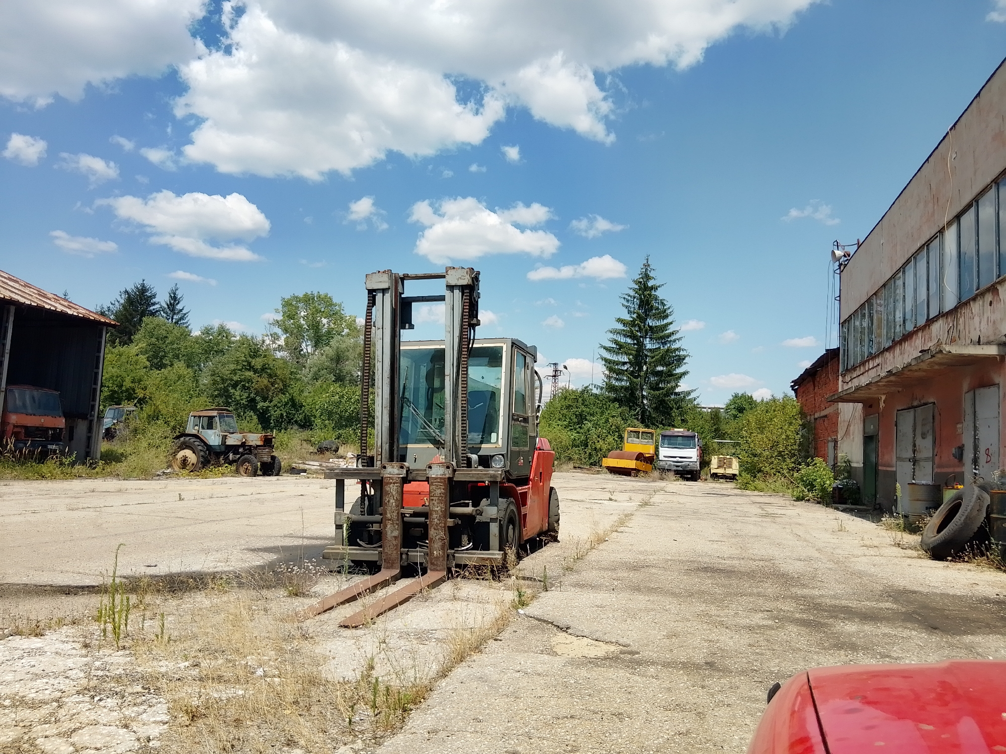In an outdoor industrial area, under a bright blue sky with scattered white clouds, a red and gray forklift is parked on a cracked concrete surface. The forklift has a metal mast and two forks. To the left of the forklift, a rusty tractor is visible, partially obscured by a building with a corrugated metal roof. In the background, trees and other industrial vehicles, including a yellow road roller and a white truck, are present. The scene appears to be a storage or maintenance yard, possibly in a rural or semi-industrial part of the city.