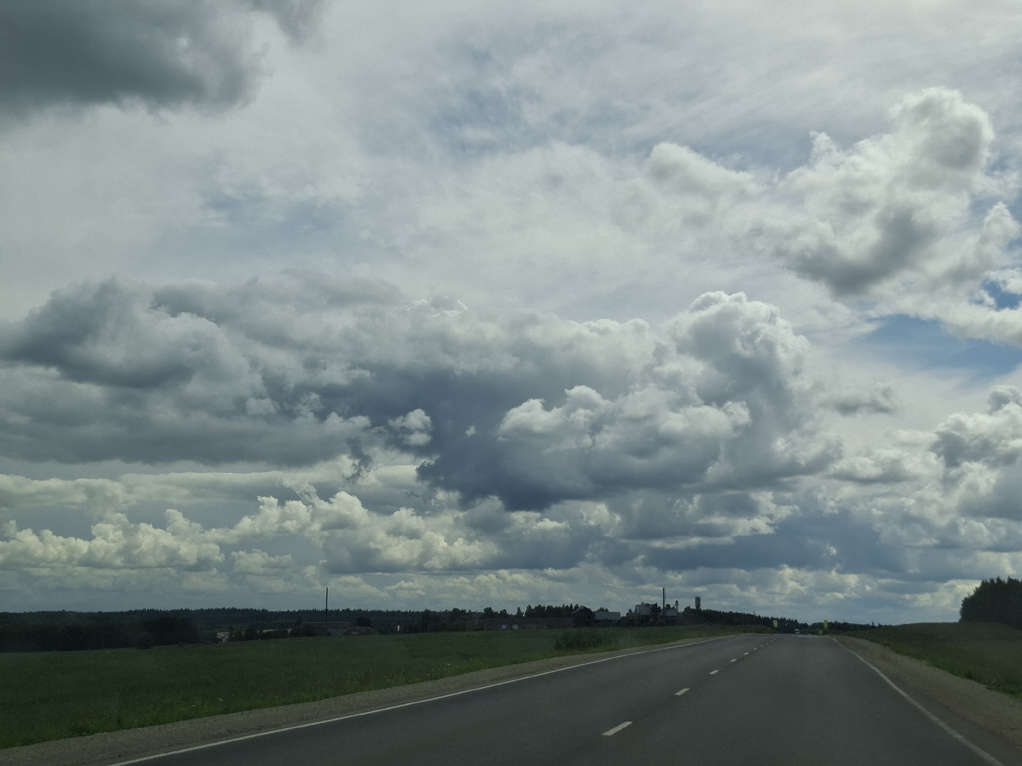 The image shows a paved road stretching into the distance through a rural landscape. On the left side of the road is a green field. On the right side of the road, the terrain slopes upwards and is covered with grass. In the distance, there are trees forming a dense forest line along the horizon. Above the landscape, the sky is filled with dramatic, heavy clouds in varying shades of gray and white, suggesting an overcast or possibly stormy weather condition. A few low-lying buildings and structures, possibly an industrial facility or farm complex, are visible in the mid-ground on the left side, nestled amongst the trees. The road itself has white dashed lines indicating lanes. The overall impression is a serene, yet somewhat imposing, natural scene with man-made elements present but not dominating the landscape.