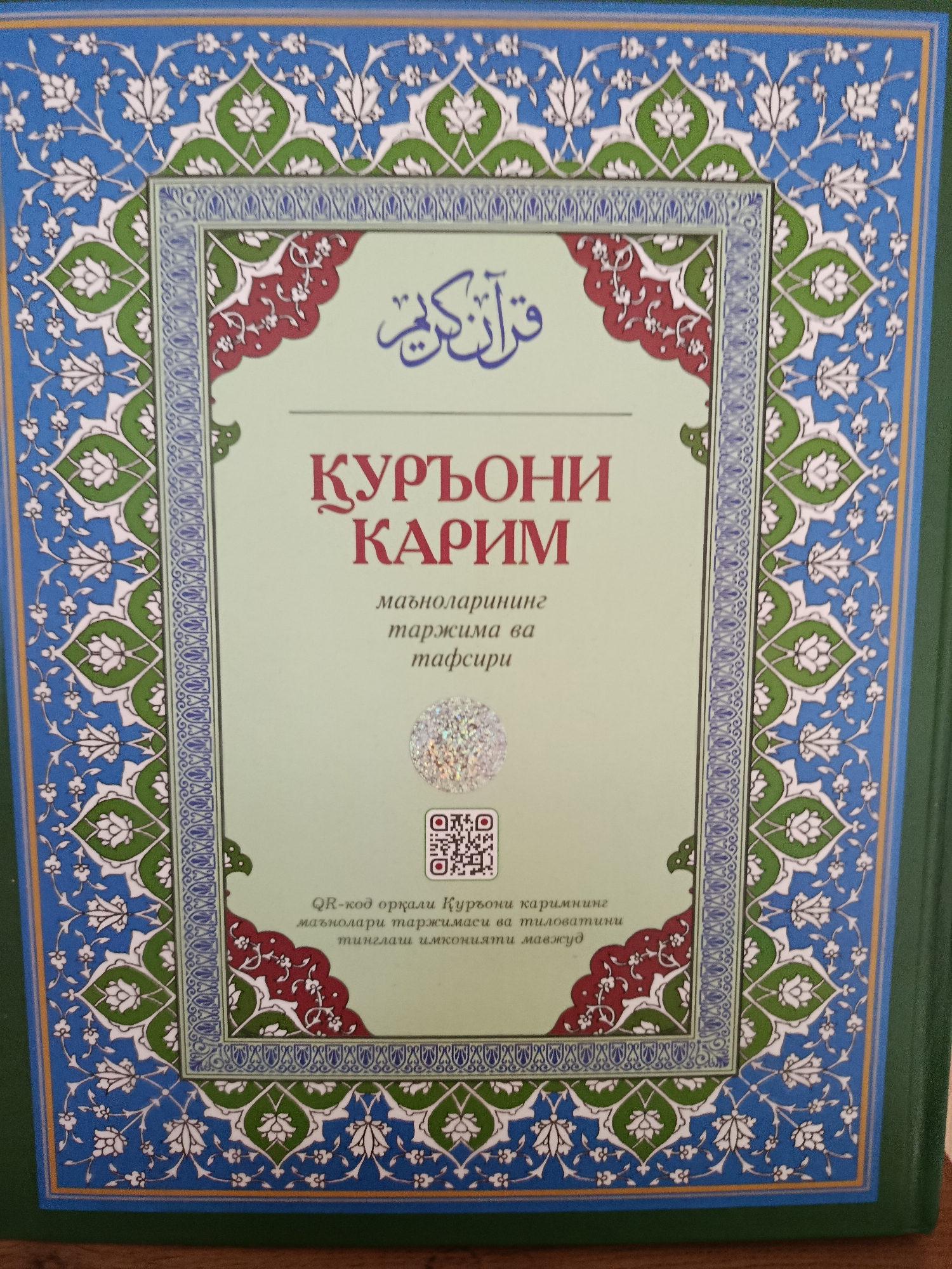 The image shows the cover of a book, likely a religious text, with intricate floral and geometric patterns. The predominant colors are blue, green, and white, with red accents in the central design. The text on the cover appears to be in Arabic script at the top and in a Latin-based alphabet below, possibly Uzbek. The book is displayed on a wooden surface. Based on the language of the text and the cultural context suggested by the design, this book could be found in Tashkent, Uzbekistan, a major cultural and religious center in Central Asia.