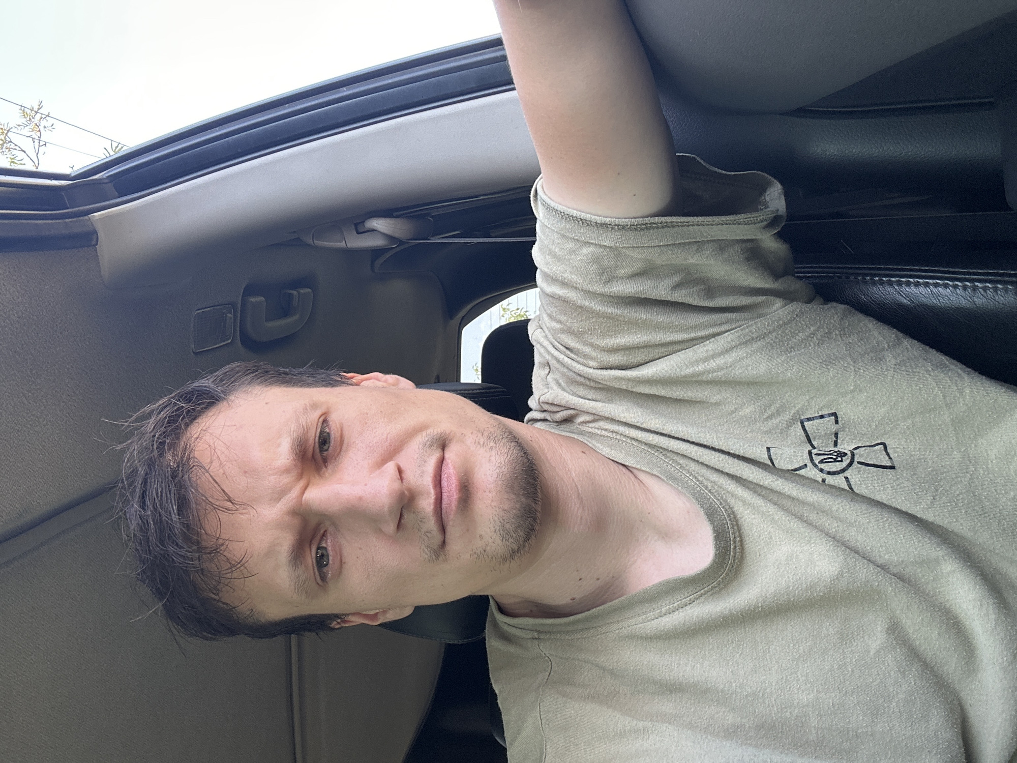 A man is sitting in the driver's seat of a car, looking at the camera. He has short, dark hair, is wearing a light brown t-shirt with a black symbol on the left chest, and appears to be in his late 20s or early 30s. The interior of the car is visible, including the headliner, door panel, and a seatbelt. The lighting suggests it is daytime, and the outdoor environment is partially visible through the car window. The man's facial expression is a slight grimace or frown, with furrowed eyebrows. His right arm is resting on the door panel. The symbol on his shirt is a stylized cross with a trident in the center, which is the national coat of arms of Ukraine. The image was taken in the city of Makiv, Ukraine.