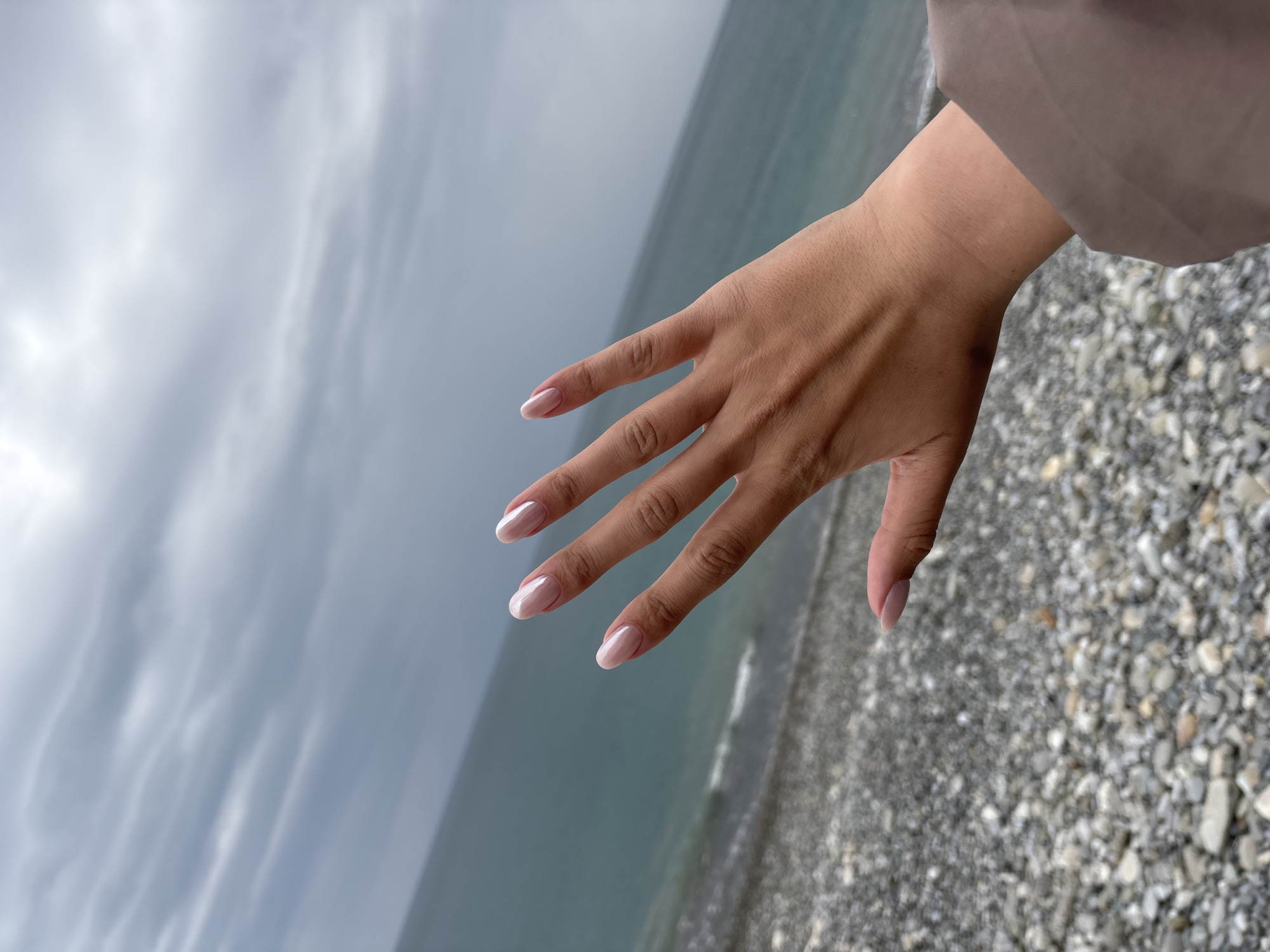A photograph displays a person's hand, positioned against a backdrop of the sea and a stony beach. The hand features neatly manicured nails with a classic French tip design. The person is wearing a light brown colored top with part of the sleeve visible near their wrist. The sea appears to be calm, reflecting the overcast sky above. The beach is composed of small, grey stones.
The image's origin is traced to Sochi, Russia.
