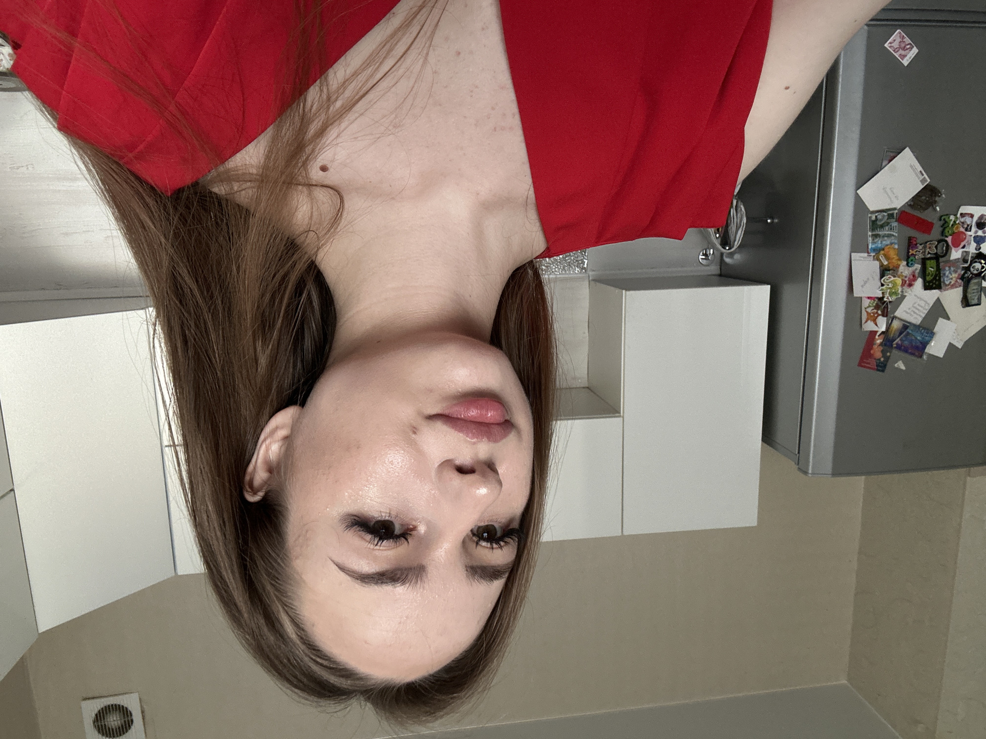 A young woman with long brown hair, wearing a red top, poses for a selfie in what appears to be a kitchen. She is looking slightly to her left, with her mouth slightly open. The background includes a gray refrigerator covered with various magnets and photos, white kitchen cabinets, and a vent on the ceiling. The lighting is soft and natural, suggesting the photo was taken during the day. The scene is casual and domestic, with no other people or significant activity visible.