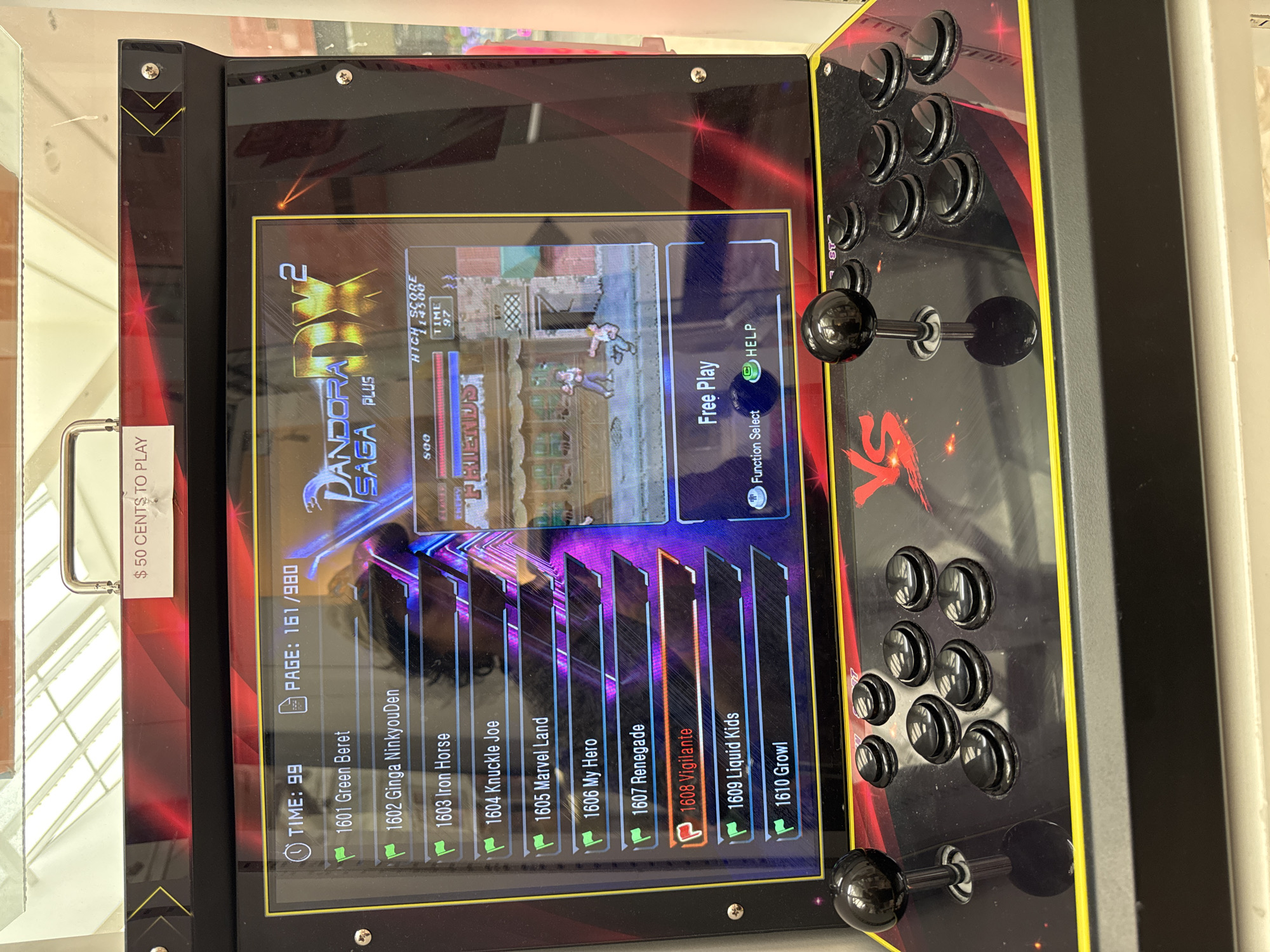 The image shows an arcade cabinet with a digital display and a control panel. The display shows a game menu for "Pandora Saga DX Plus" with a list of game titles and a game in progress. The control panel features two joysticks and several buttons. A sign at the top of the cabinet reads "$50 CENTS TO PLAY". The arcade cabinet is positioned in front of a retail display with fluorescent lighting overhead. The scene appears to be indoors, likely in a store or arcade, and is located in El Paso, United States of America. There are no visible people in the immediate vicinity of the arcade cabinet. The overall impression is that of a typical arcade setup.