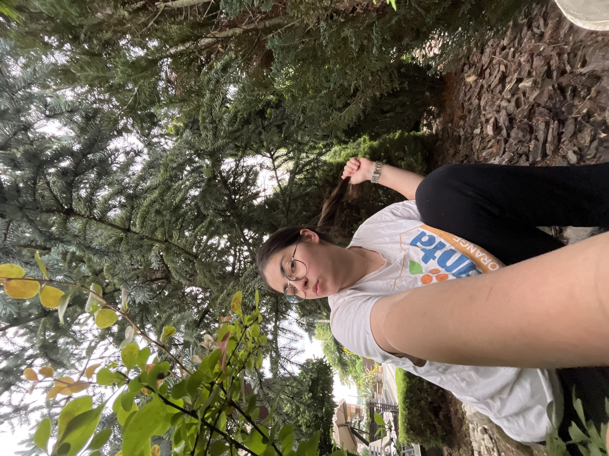 A young woman with glasses and her hair in a ponytail is taking a selfie outdoors in a backyard setting. She is wearing a white t-shirt with orange and blue lettering and black pants. She is holding a strand of her hair in her right hand. The background is filled with dense evergreen trees. In the distance, outdoor furniture and landscaping are visible, suggesting a residential area. The lighting indicates it is daytime. There is no discernible indication of the city of Koroviia, Ukraine.