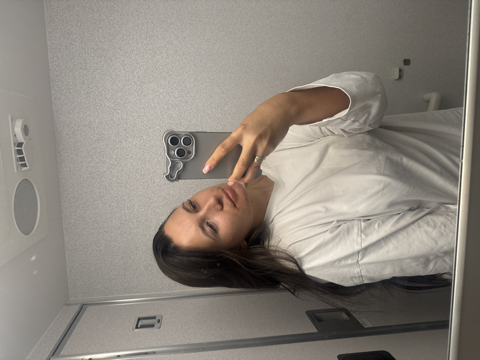 A person is captured in a mirror selfie, holding a smartphone to take their own picture. The image is oriented vertically, as if the camera was rotated 90 degrees clockwise from a standard landscape shot.
The subject is a woman with long, dark brown hair, which is parted on her left side and cascades over her left shoulder and down her back. She has a fair complexion with some minor blemishes visible, and her eyebrows are dark. Her eyes are looking directly towards the phone, and her lips are slightly parted, revealing a neutral or contemplative expression. She appears to be wearing a plain, light-colored (likely white or off-white) long-sleeved t-shirt or top, which is loose-fitting and shows some wrinkles.
Her right hand holds a gray smartphone, which appears to be a modern iPhone model with a prominent triple-lens camera module. The phone is encased in a distinctive, light gray, irregularly shaped case that features a wavy or organic design element on the left side and a clear cut-out for the cameras. Her fingernails are painted a light, subtle color, possibly pink or nude. On her right ring finger, she wears a stack of at least two thin, metallic rings.
The setting appears to be an interior space with light gray, speckled or textured walls, possibly resembling a small room like an airplane or train lavatory, or a compact cabin space. In the upper left portion of the reflection, a white vent or air conditioning unit is visible, with a round speaker-like grill directly beneath it. The bottom left of the reflection shows what looks like a metallic handle or latch, possibly part of a door. The right edge of the mirror frame is visible, appearing white or light-colored. The lighting is even, suggesting indoor artificial light. No discernible text is visible anywhere in the image.