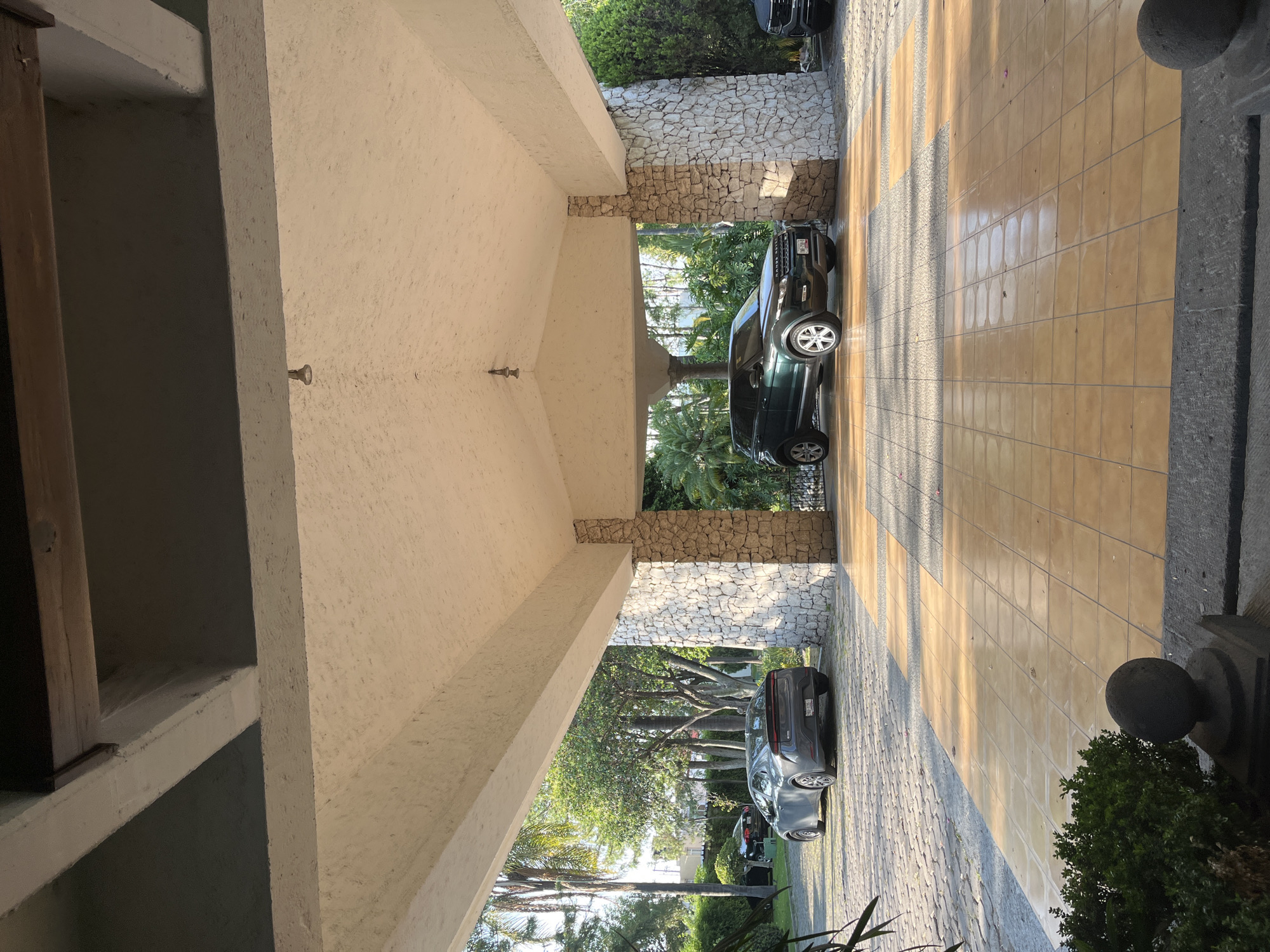 The image shows a covered driveway with two cars parked on a tile surface in San Bernardino Tlaxcalancingo, Mexico. The driveway is framed by stone walls and beige textured supports leading to a building on the left. The right side of the driveway is lined with a stone border and greenery. The cars are a dark green SUV and a gray sedan. Beyond the driveway, trees and sky are visible. The time of day appears to be mid-day based on the brightness of the sunlight.