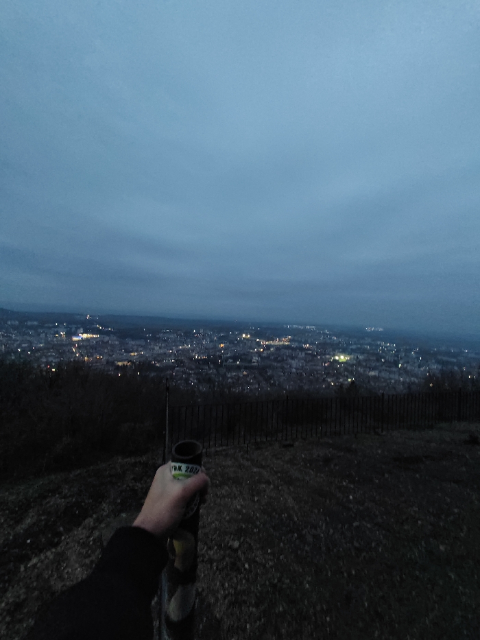 The image captures an elevated nighttime view overlooking the city of Shumen, Bulgaria, under an overcast, dark blue-grey sky. In the foreground, a human left arm, clad in dark long-sleeved clothing, holds a vertical, dark cylindrical object. A light green label reading "MK 200" is visible on the object, which appears to be a can or bottle cooler, partially revealing a yellow item inside or wrapped around it. A dark metal fence spans horizontally across the mid-ground, separating the viewer's immediate position on a rocky, dark ground surface from the distant city. The city of Shumen is illuminated by numerous scattered streetlights and building lights, creating a glowing urban expanse against the dark background.