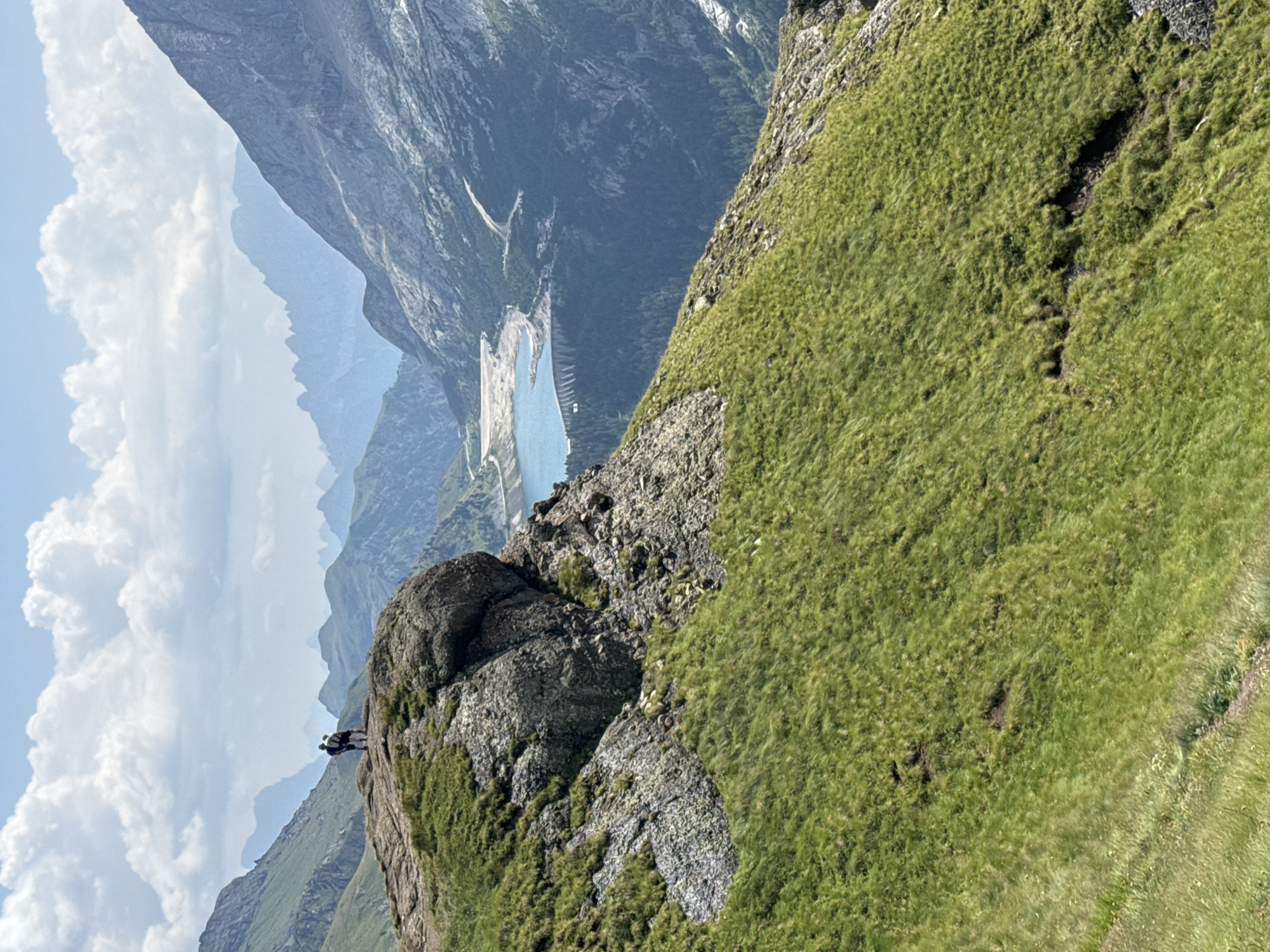 The image depicts a panoramic view of the Dolomites mountain range near Canazei, Italy. In the foreground, two figures stand on a rocky outcrop covered in lush green grass. They are facing away from the camera, looking out at the vast landscape. Behind them, a scenic valley unfolds, featuring a vibrant blue lake and a prominent dam. The mountains in the distance are characterized by their jagged peaks and rocky slopes, shrouded in a light haze. The sky is partly cloudy, with soft white clouds drifting against a clear blue backdrop. The overall impression is one of natural beauty and serenity.