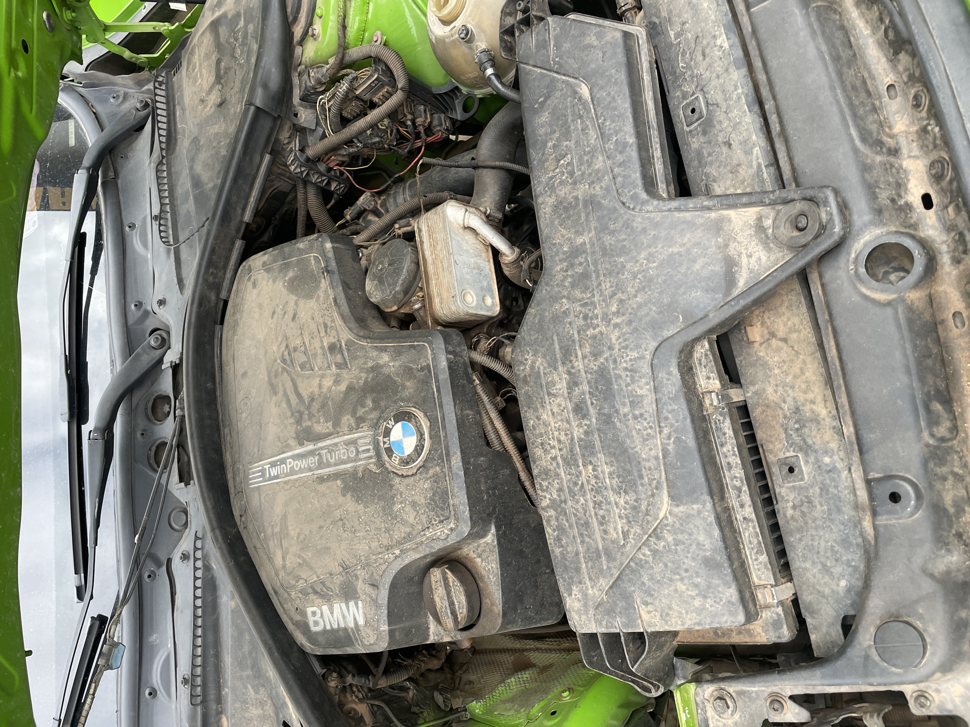 The image shows a dusty BMW engine bay. The engine is a "TwinPower Turbo" model. The car's body is painted green. The location and city of Hayin Banki, Nigeria, are not discernible from the image. There are no visible people or actions in the scene. The primary subject is the car engine. The environment is a typical engine bay interior.