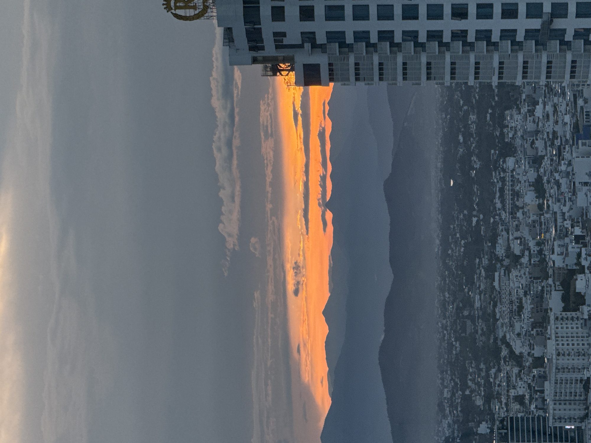 This is a high-angle, wide shot of a city skyline at dusk, with a dramatic sunset in the background. The sky is filled with layers of clouds, with a vibrant band of orange and yellow light radiating from the horizon where the sun is setting behind a range of silhouetted mountains. In the foreground, a tall, modern white skyscraper dominates the right side of the frame. The building features a repeating pattern of windows and balconies, and at its peak, a golden emblem, possibly a logo, is visible. Below the skyscraper, the city stretches out into the distance, a dense collection of buildings with rooftops and varying architectural styles. The cityscape appears somewhat hazy, suggesting either distance or atmospheric conditions. The mountains in the background are layered and fade into a bluish haze, with the sunset light catching their upper edges. The overall impression is one of urban development against a natural, colorful sky. The image does not show any people.