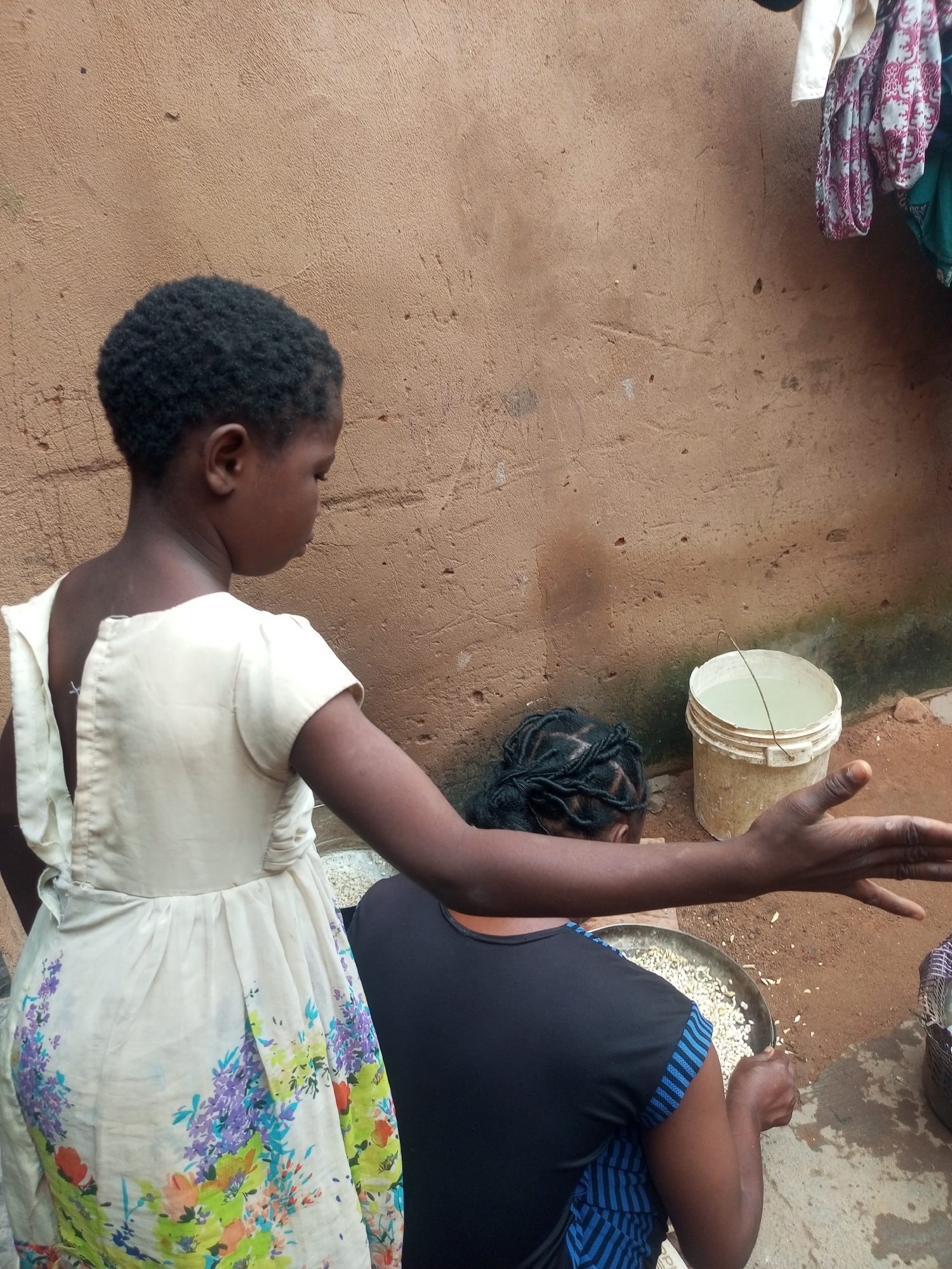 Two young girls are engaged in an outdoor activity in a dusty, outdoor area with a rough, textured brown wall as the backdrop. The girl in the foreground, wearing a light-colored dress with a floral print, stands with her back to the camera, her right arm extended outwards as if gesturing. Her hair is short and dark. Behind her, a second girl, her back also turned, is seated and focused on a large, shallow metal pan filled with what appears to be grains or seeds. She has dark hair styled in braids. To the right of the frame, a pale plastic bucket filled with water is visible. Clothing, possibly laundry, is hanging on the wall in the upper right corner. The ground is composed of reddish-brown earth. The lighting suggests it is daytime. The setting appears to be a residential or communal outdoor space, possibly in Gombe, Nigeria, given the characteristic brown, rough-textured walls often found in such regions.