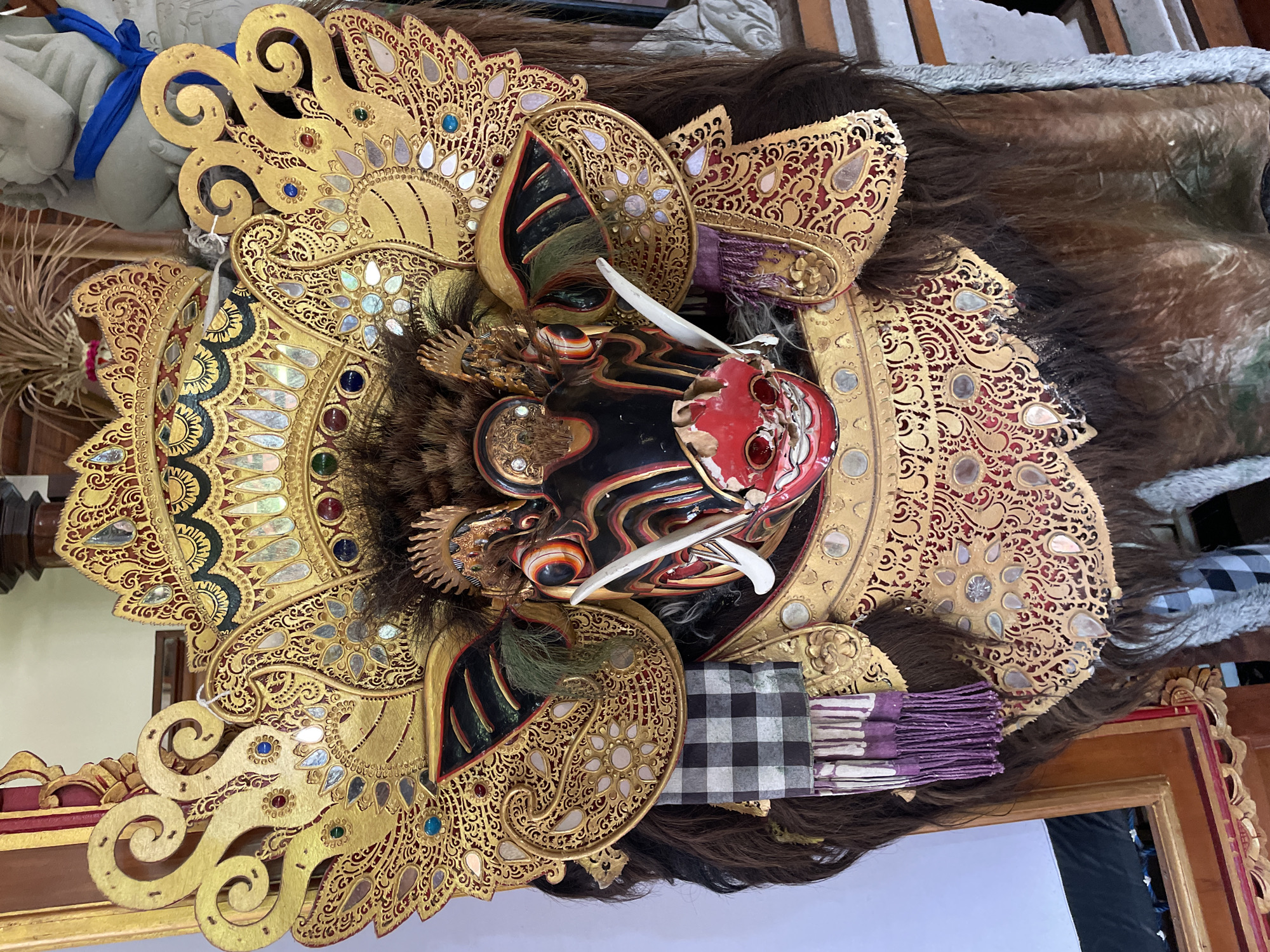 An ornate, elaborately decorated mask is displayed prominently in the center of the frame. The mask is predominantly gold and black, with red accents on the face and mouth. It features large, stylized ears and a headdress adorned with intricate patterns, small mirrors, and colorful gemstones. Long, dark hair emanates from the sides and bottom of the mask. A black and white checkered fabric with purple tassels is draped over the left side of the mask. The mask is situated in an indoor setting, with hints of wooden furniture and a light-colored wall in the background. There are no people or animals visible in the image. The lighting suggests an ambient indoor environment.