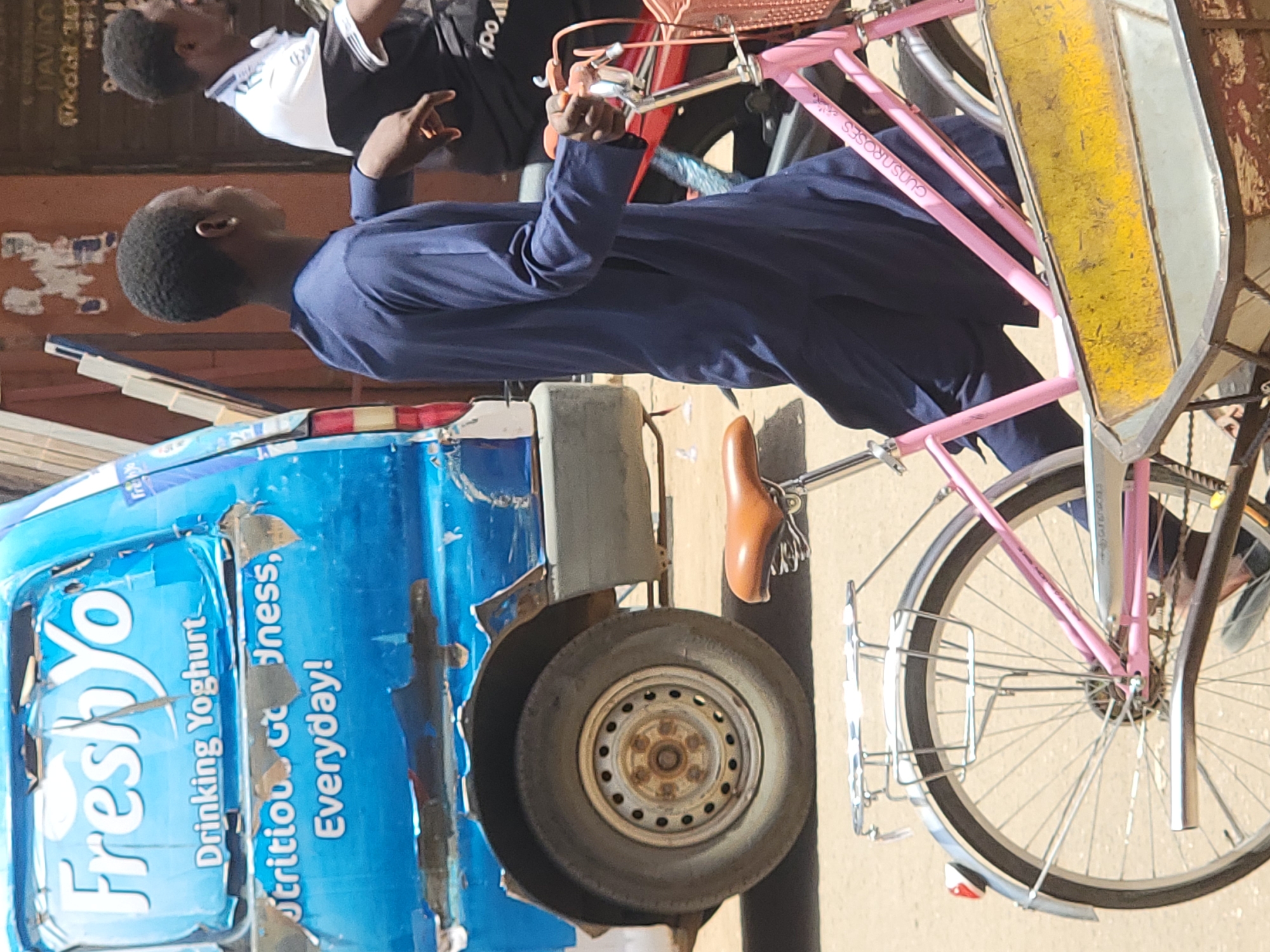 The image, rotated 90 degrees clockwise, captures a vibrant street scene in Maiburiji, Nigeria, under bright daylight. In the foreground, a young man with short, dark hair is depicted in profile, wearing a dark blue, long-sleeved garment that appears to be a traditional or work uniform. He is standing next to a bicycle and appears to be holding its handlebars with his right hand.
The bicycle has a distinctive pink frame, a brown leather-look saddle, and silver handlebars. The words "GUNS N' ROSES" are visible in white lettering on the pink frame. A wire basket or carrier is attached to the front of the bicycle.
To the left of the man and bicycle, a blue, somewhat weathered vehicle or cart is prominent. Its side displays legible text, advertising "FreshYo Drinking Yoghurt" and "Nutritious Goodness, Everyday!" The blue surface shows signs of wear and peeling. A single wheel of this vehicle/cart is visible, suggesting it might be a three-wheeled delivery vehicle or a modified cart.
In the background, a second person is partially visible, wearing a black and white striped top. The setting suggests an outdoor market or street area with other structures in the blurred background. The overall impression is one of daily life in an urban or semi-urban African environment.