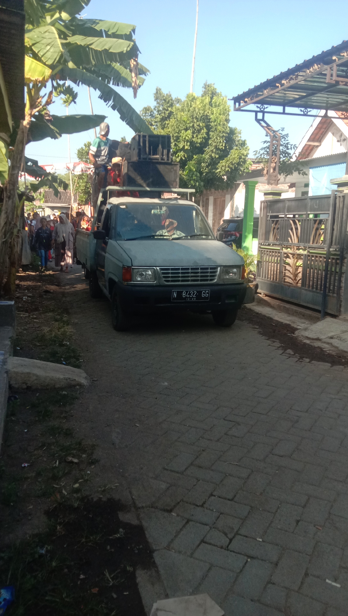 An outdoor, slightly low-angle shot shows a light gray pickup truck parked on a paved street lined with brick. The truck's license plate reads "N 8432 GG." The truck bed is equipped with several large speakers. A person wearing a cap and a green and white shirt stands on top of the speakers, appearing to be operating them. To the left of the truck, a group of people, some wearing traditional clothing, walk along a dirt path. Banana trees with large leaves are visible in the upper left corner. In the background, a house with a covered carport and a metal gate can be seen. The sky is clear and blue. The scene is located in Ngempit, Indonesia.