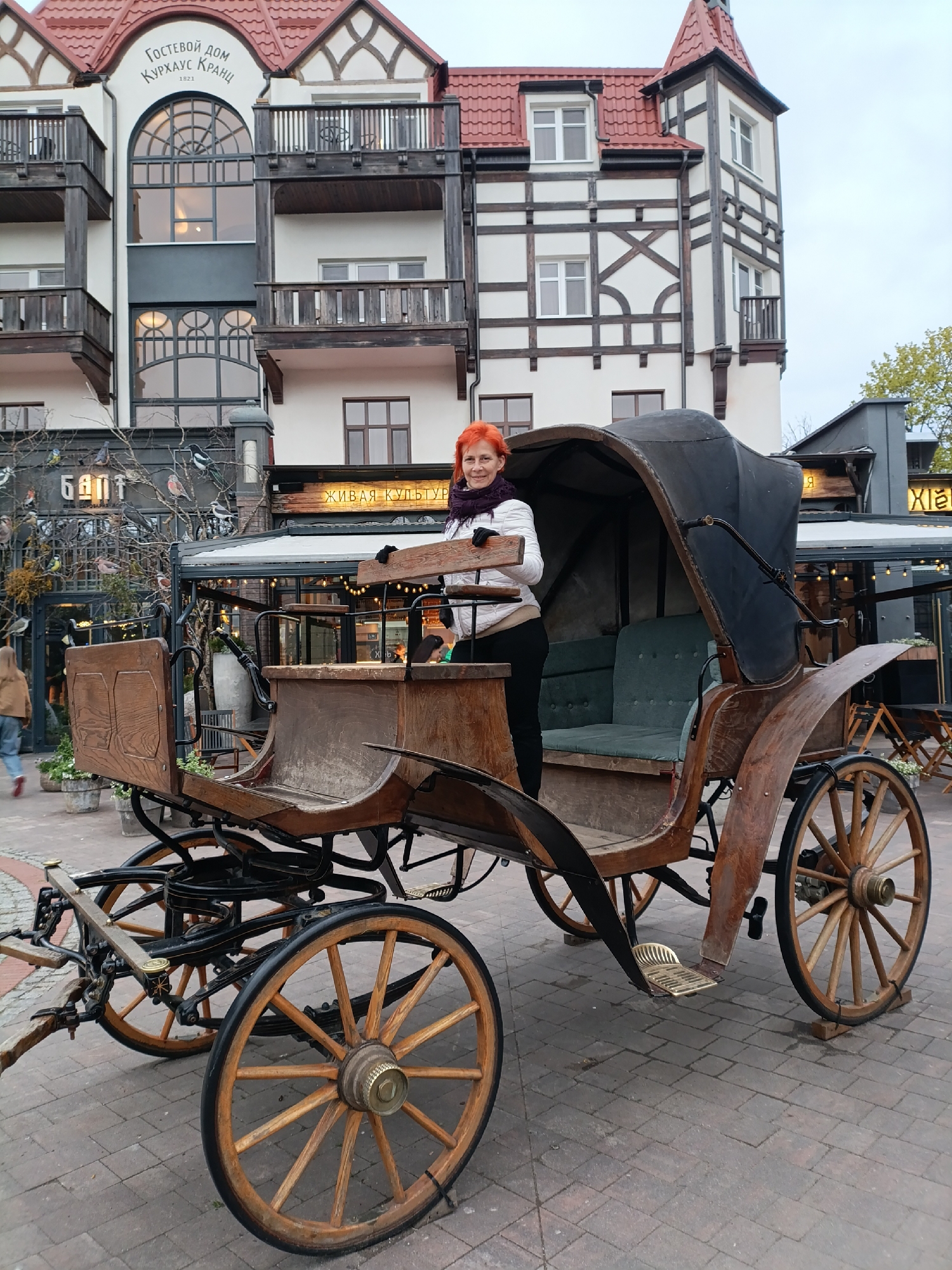 A full shot captures a woman standing in a horse-drawn carriage in Zelenogradsk, Russia. The woman, with short red hair, wears a white jacket and black pants. The carriage, made of dark wood with black accents, has a partially covered seat. Behind the carriage are buildings with European architectural styles, including timber framing. The ground is paved with bricks. Signs in Russian are visible on the buildings in the background.
